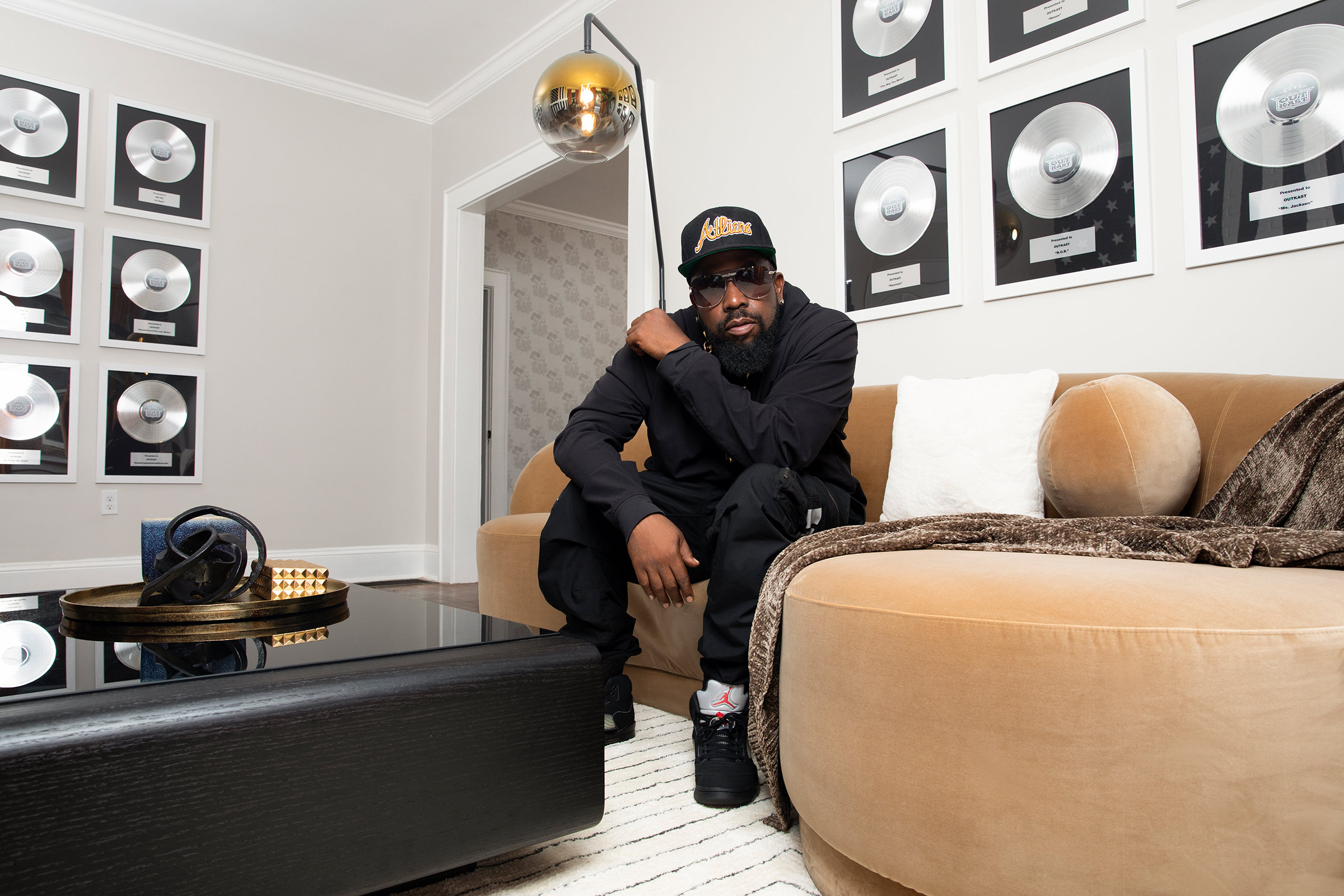 Big Boi sitting on a plush brown chair with wall of platinum records hanging on the wall behind him
