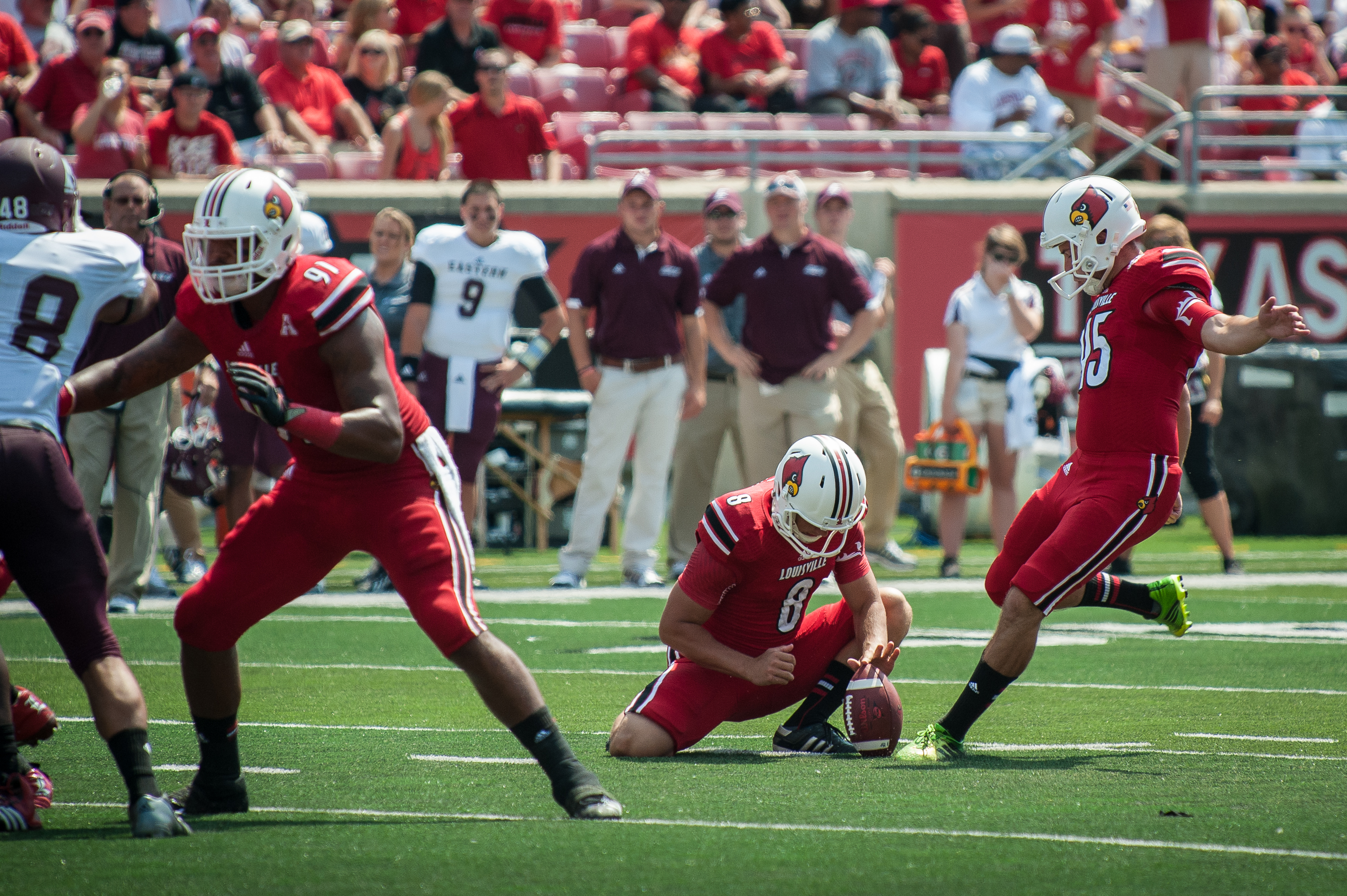 A Louisville Cardinals football player is about to kick a football with coaches, players, and fans in the stands in the background