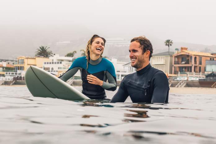 Two surfers in wet suits laugh in the water as they wait for the next big wave.