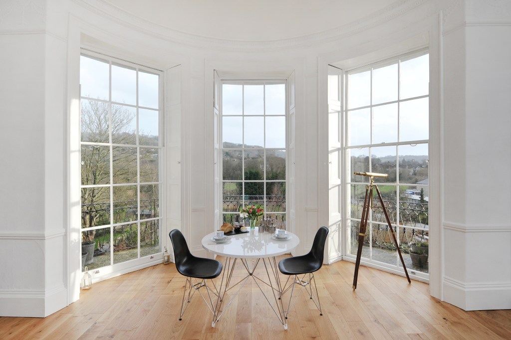 A white table and two small black chairs set against a bay window with a telescope in the background overlooking a park.