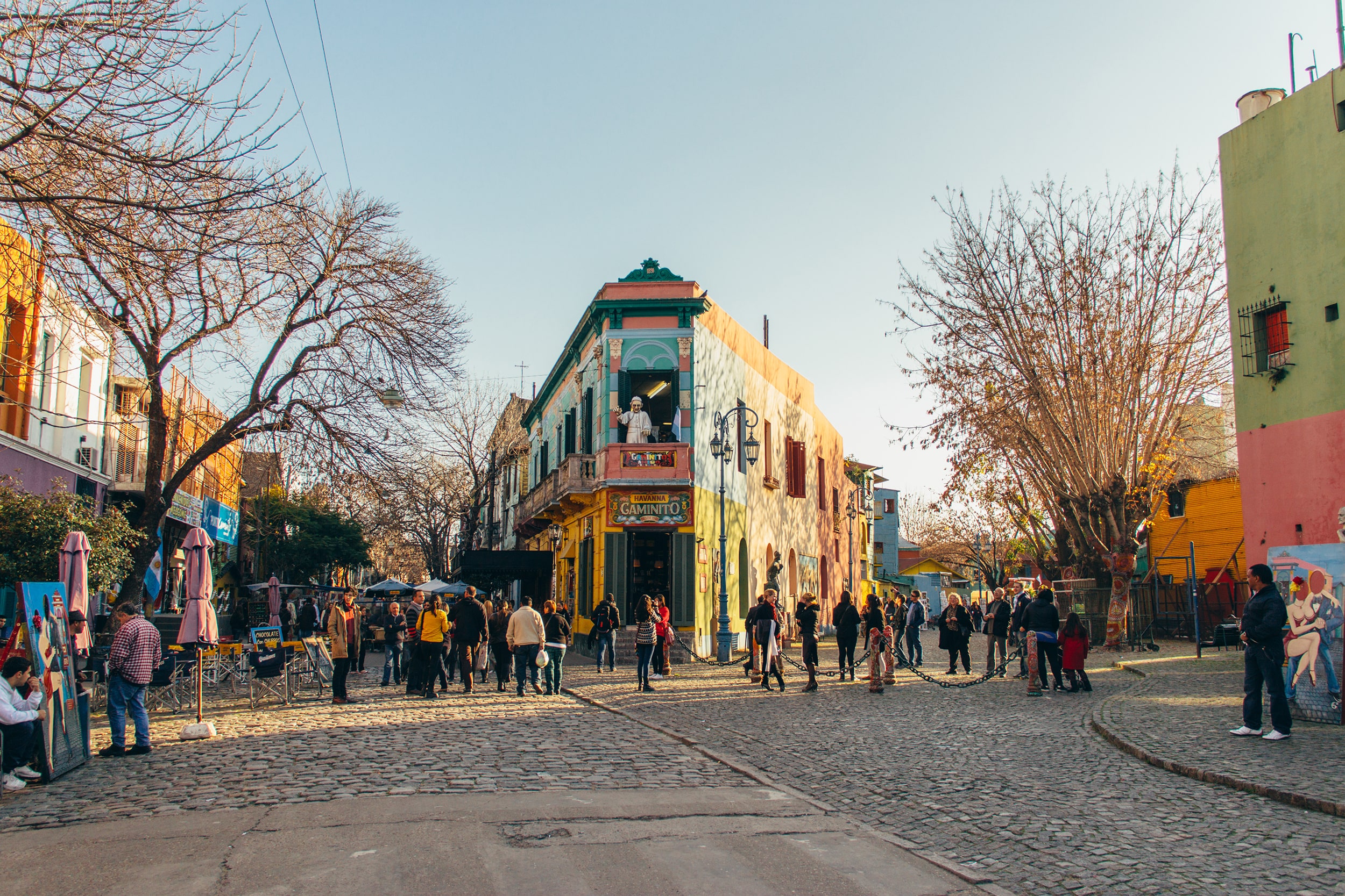 "A vibrant street scene in a colorful neighborhood, featuring a lively crowd gathered around a cobblestone square. The buildings are adorned with bright murals, and trees line the street, suggesting a warm, sunny day. People are mingling, some sitting at outdoor tables, enjoying the atmosphere."