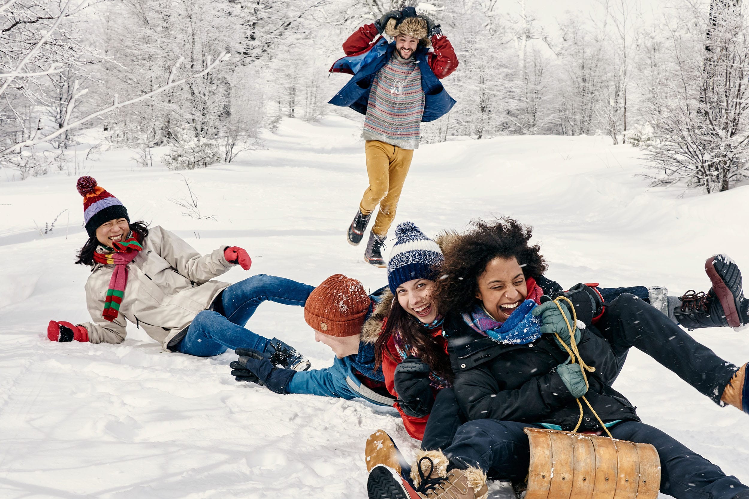 Family playing in the snow, three of them are on a sled and two more are behind, one person running and the other is lying down.
