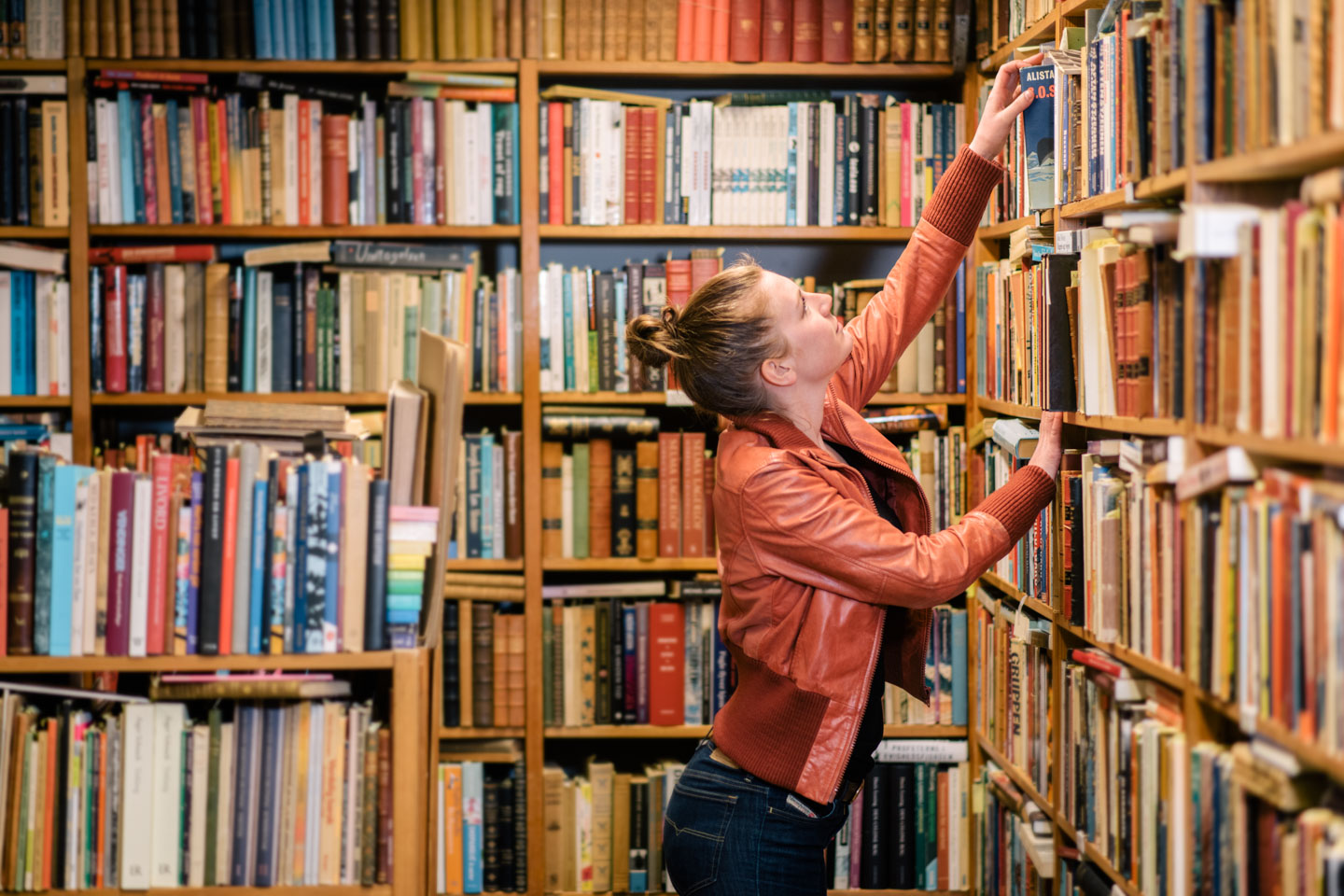 Young woman reaching to the top shelf of a bookcase in a library or bookstore.