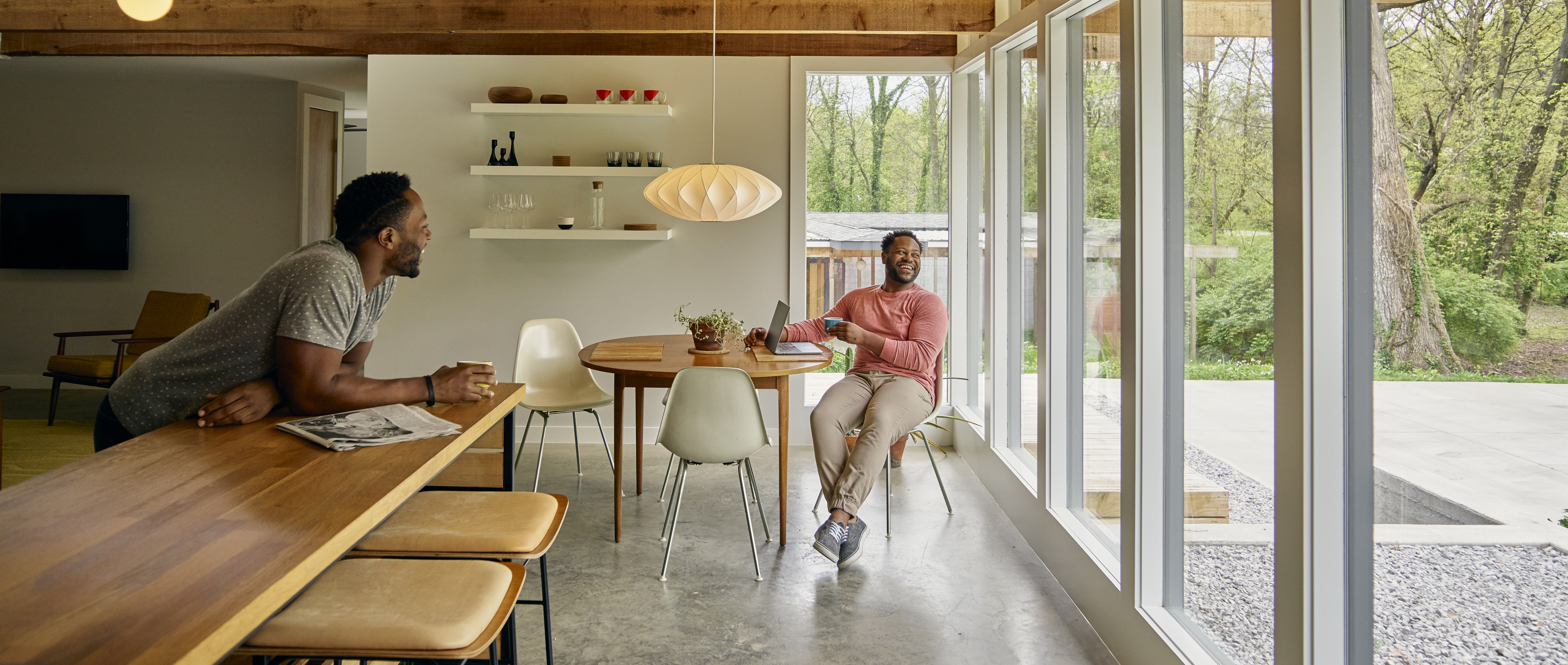 Two Black American men laughing together in a modern Airbnb listing. The sun shines through the floor to ceiling windows into the dining area.