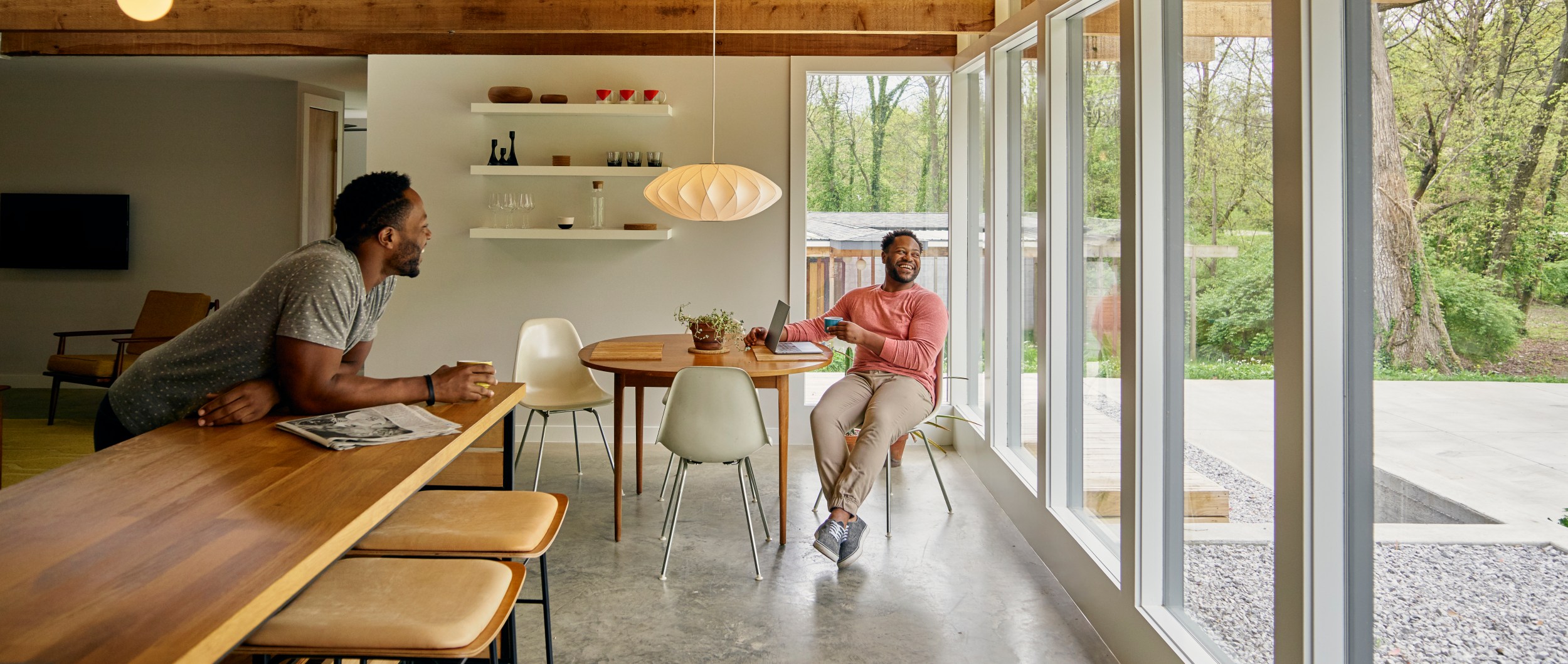 Two Black American men laughing together in a modern Airbnb listing. The sun shines through the floor to ceiling windows into the dining area.