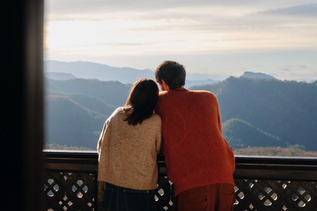 Two travelers look out from a balcony at their Airbnb listing at a beautiful mountain landscape.