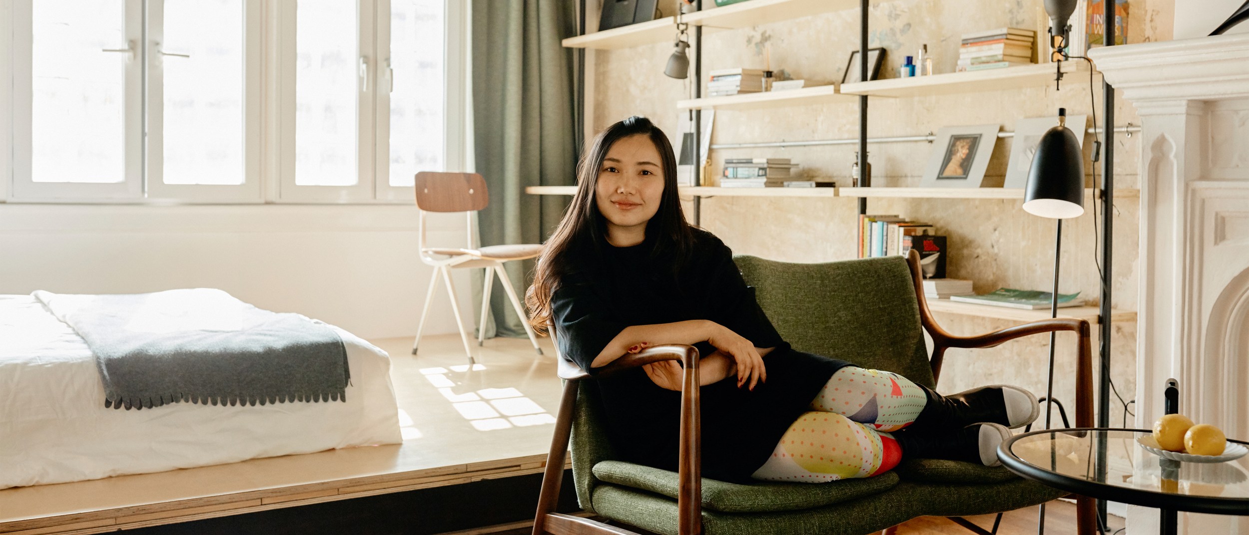 An Airbnb host in China sits on a green fabric chair in the bedroom of her listing.