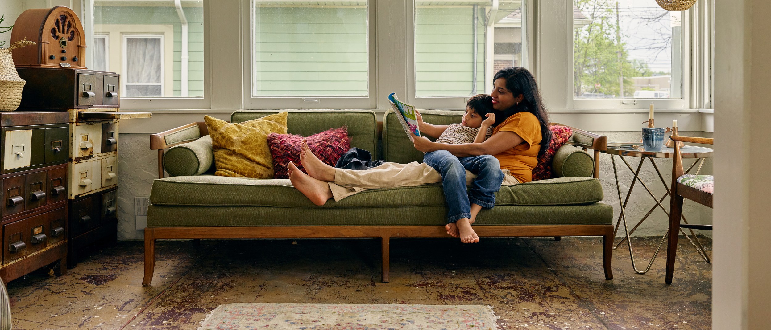 A mother reads a magazine on a vintage couch with her son in her lap while staying at an Airbnb listing.