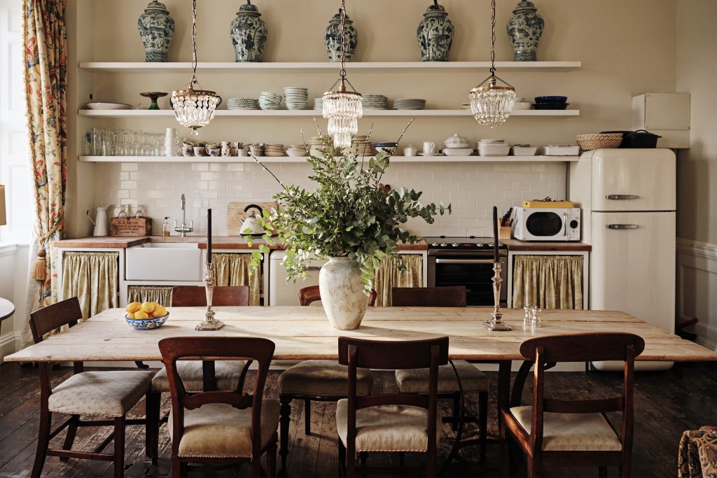 A clean and bright Airbnb dining room with 3 antique chandeliers and ha large plant in a vase at the center of a wooden table.