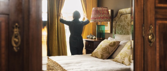A woman opens the curtains and light pours into the bedroom of her Airbnb listing.