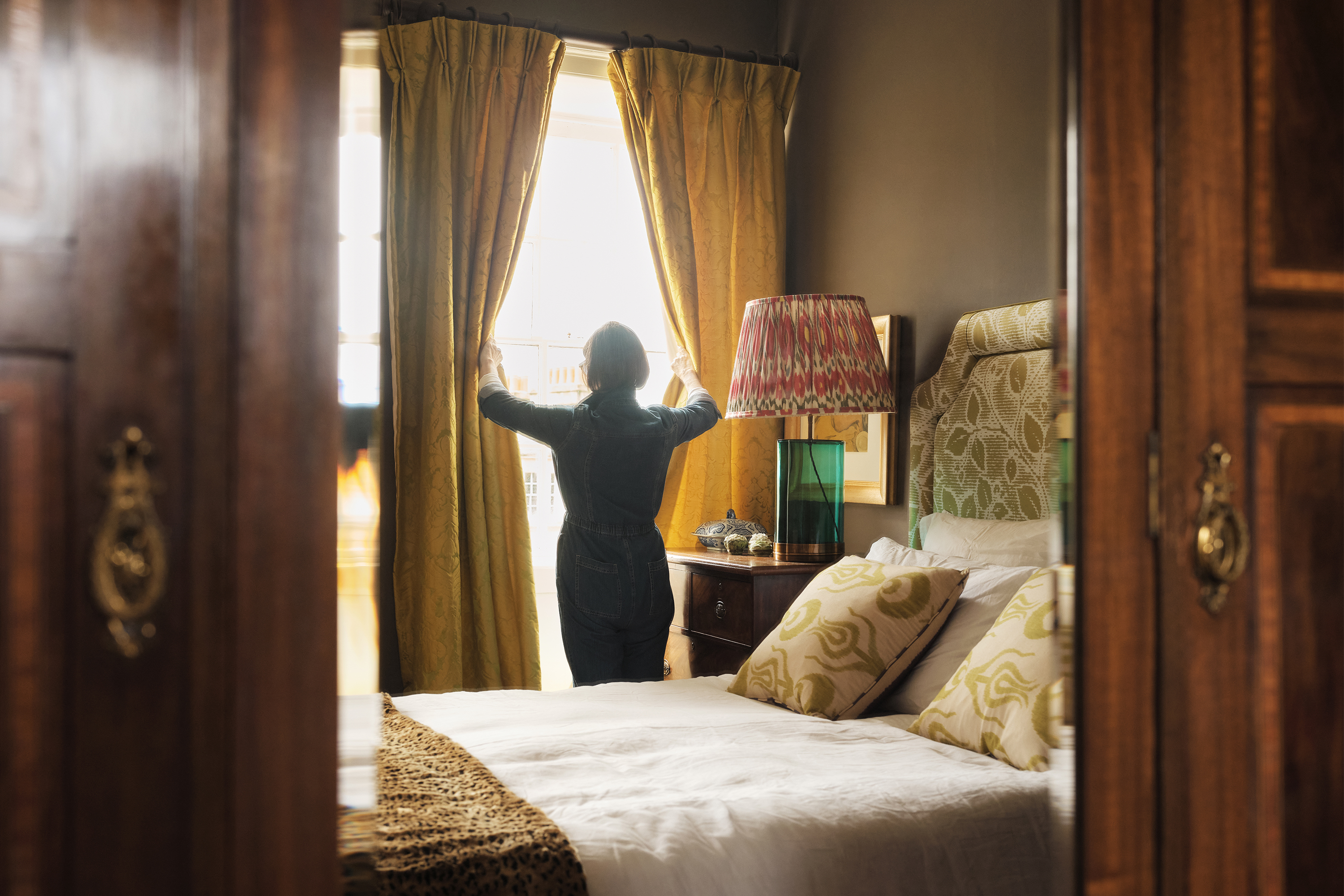A woman opens the curtains and light pours into the bedroom of her Airbnb listing.