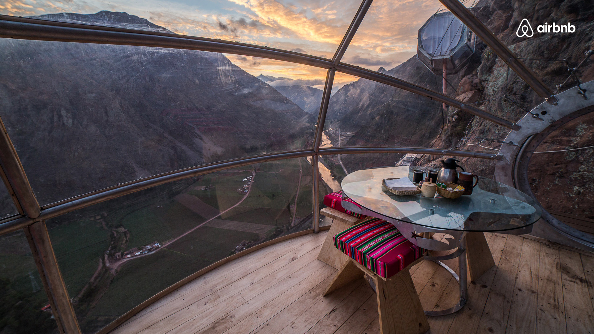 A cozy dining setup inside a glass bubble overlooking majestic mountains at dusk. The table is set with tea and snacks, surrounded by warm wooden flooring, while the panoramic view showcases a vibrant sky painted with hues of orange and blue as the sun sets behind the peaks.