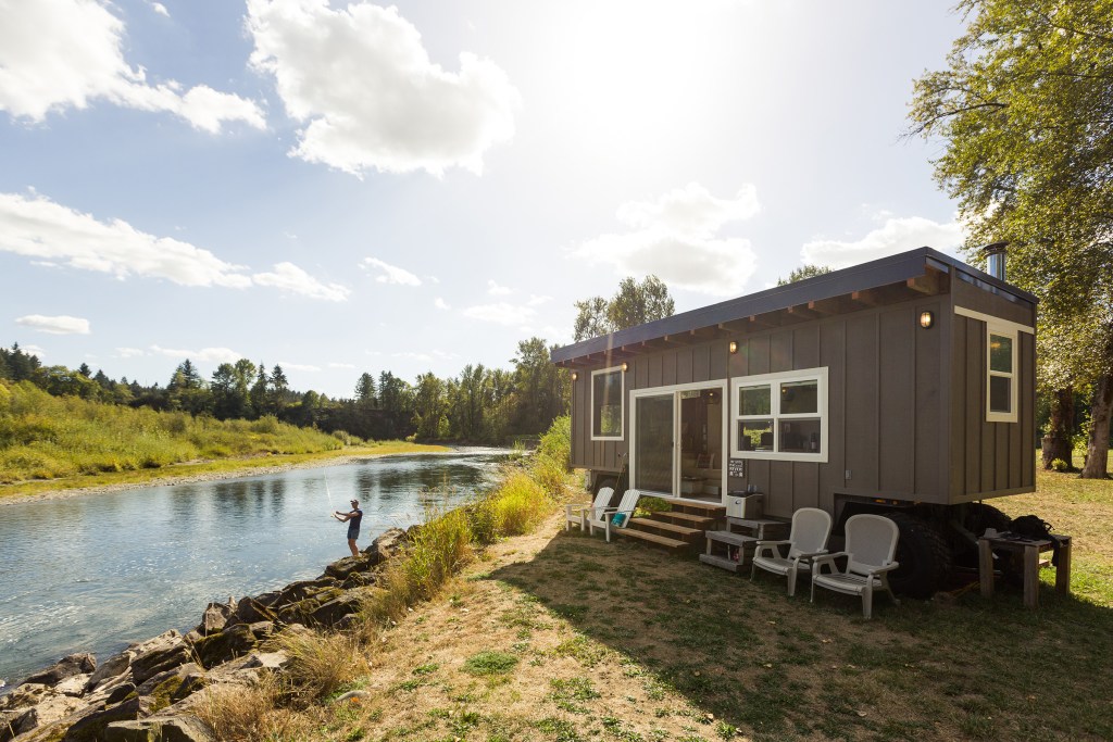 A man is fishing in a river next to the remote tiny home Airbnb listing he is staying in.