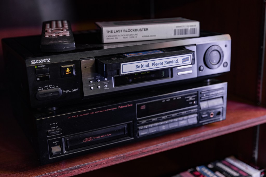A BLOCKBUSTER VHS tape is being inserted into a tape player. "Be kind. Please rewind" is printed on a label on the tape.