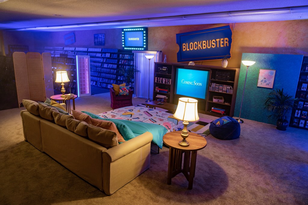 A futon with a '90s blankets is situated in front of a large TV inside the last BLOCKBUSTER store.