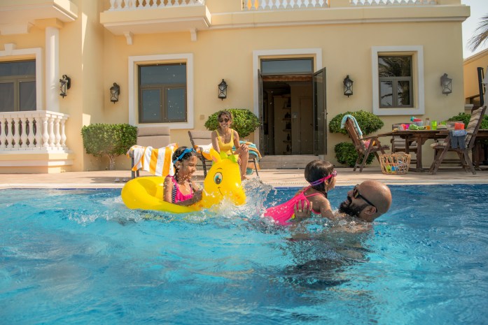 Family playing in a pool. One young girl is on a yellow floatation device, the other younger girl is being held by her father. Their mother is on the side of the pool watching.