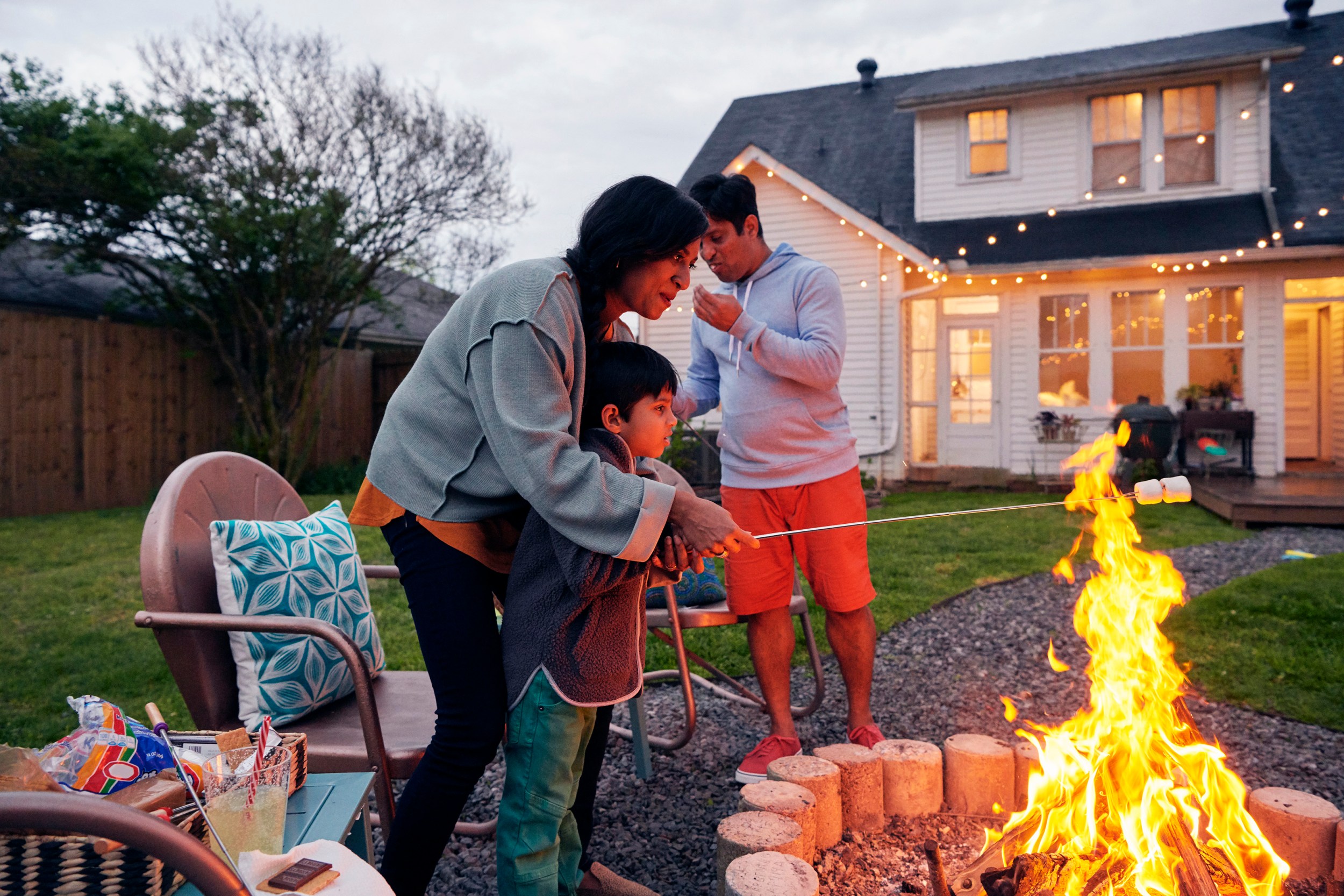Woman cooks s'moors over an open fire with her son, while a man stands in the background in front of a white house.
