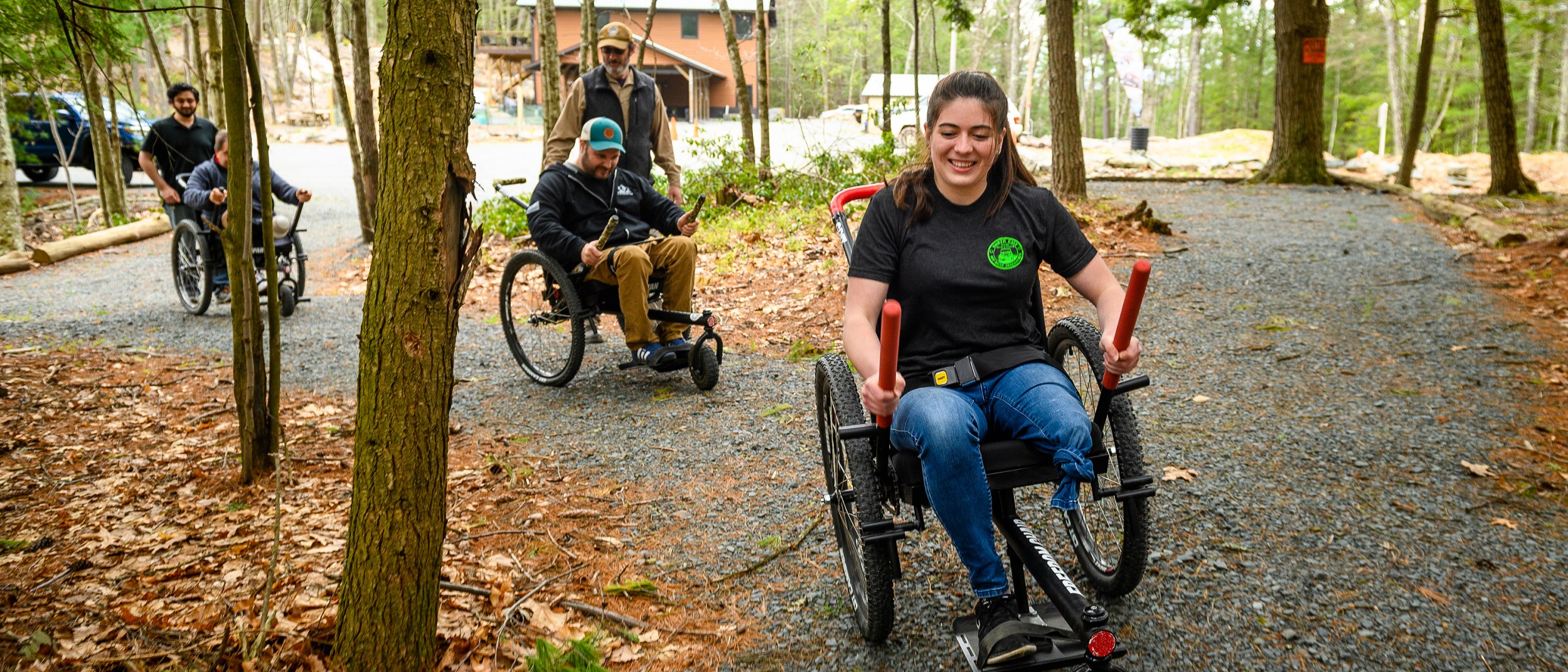 Three people using Grit Freedom Chair wheelchairs along a gravel path between some trees