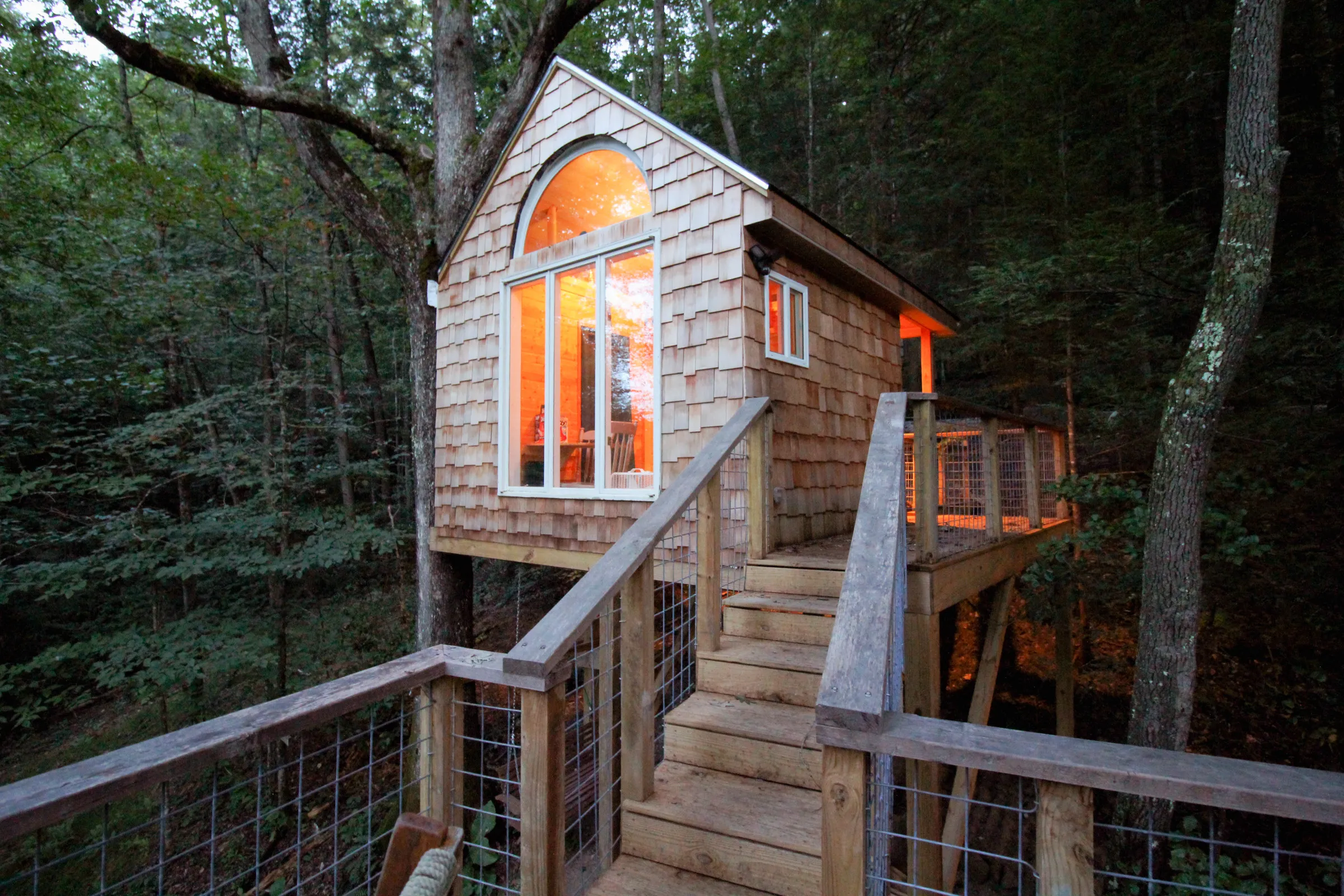 Wood shingled treehouse with staircase leading up to entry door in evening light and a glowing light coming from the window.
