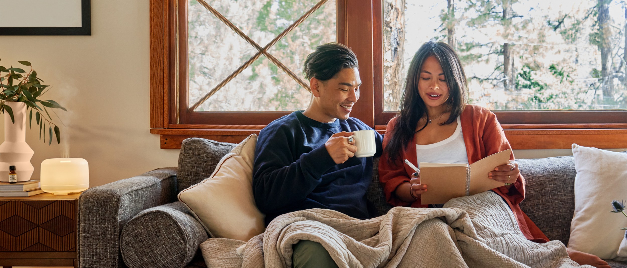 Couple sitting on the couch with a blanket and mugs, looking at a journal with an essential oil diffuser on the table next to them.