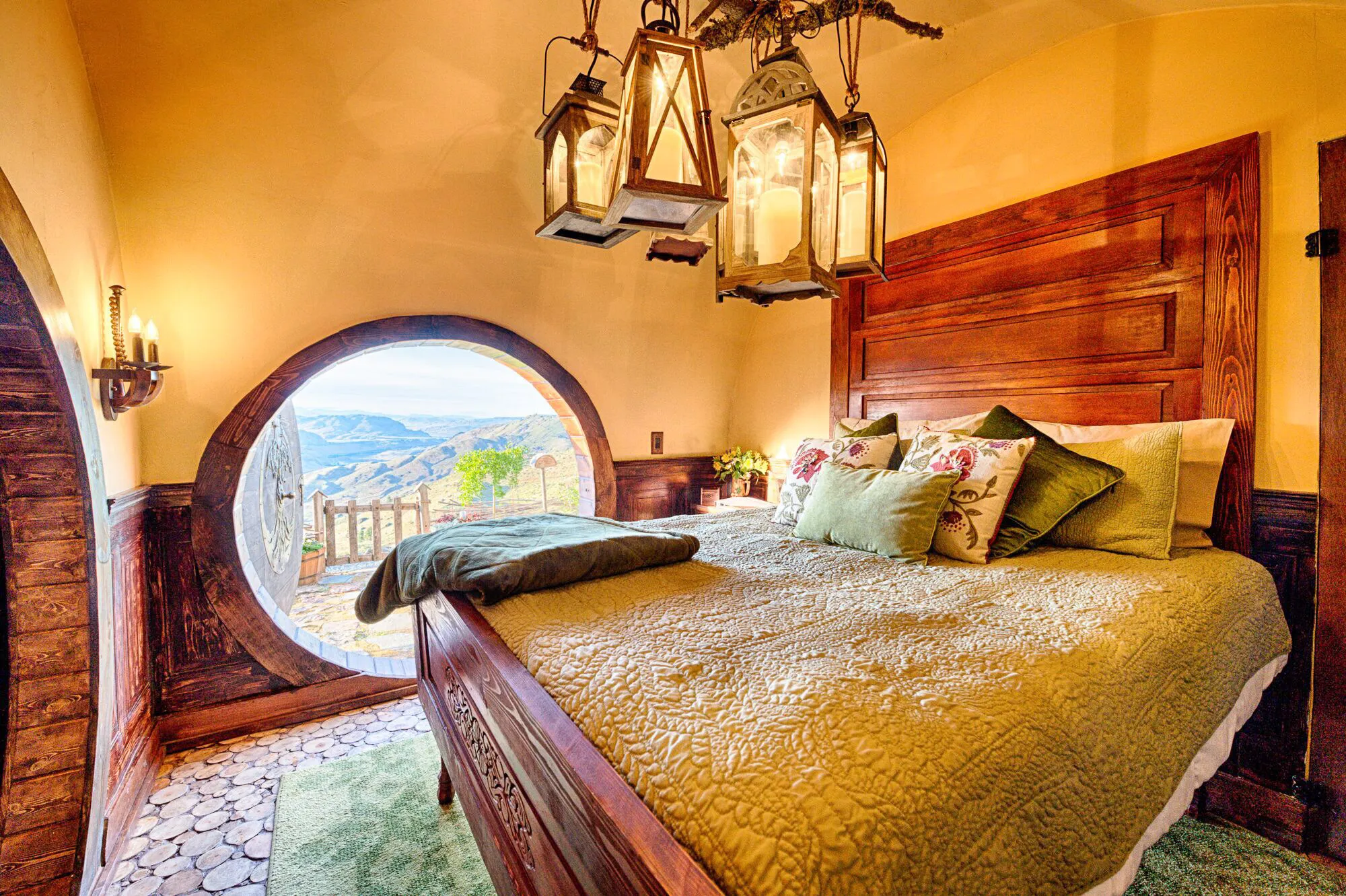 Queen sized bed with green bedding in front of a circular doorway with a rolling hillside outside.