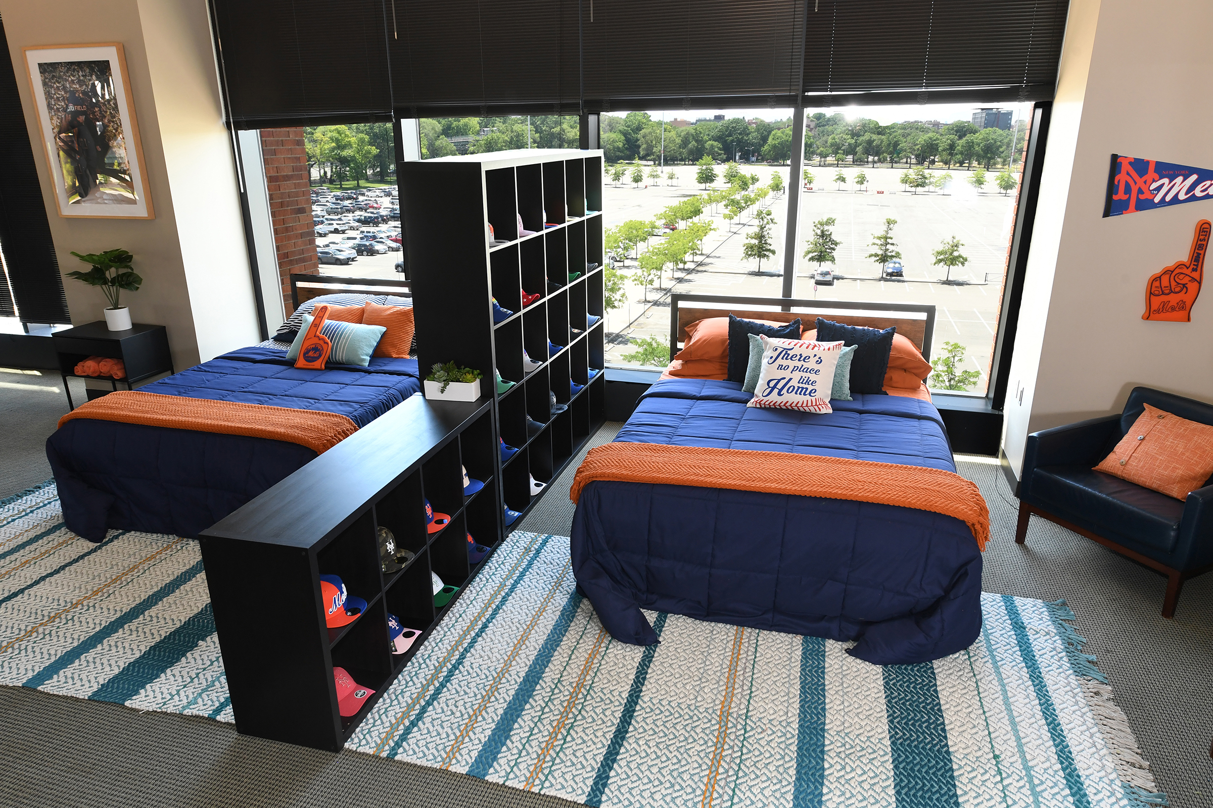 The Citi Field bedroom with a blue quit and orange throw blanket, with a bookshelf full of baseball caps in the background.