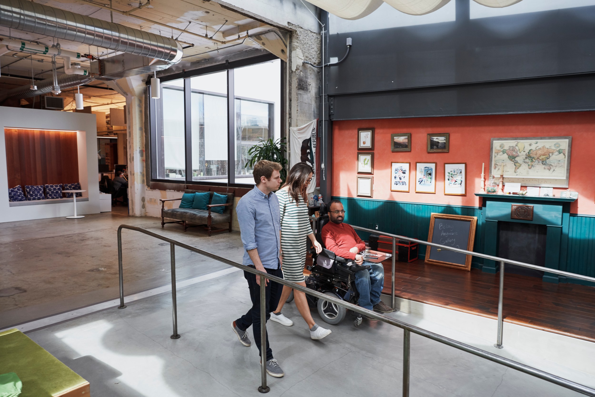 Three people are talking as they walk down a ramp in an office, including a man, a woman and a man using a wheelchair