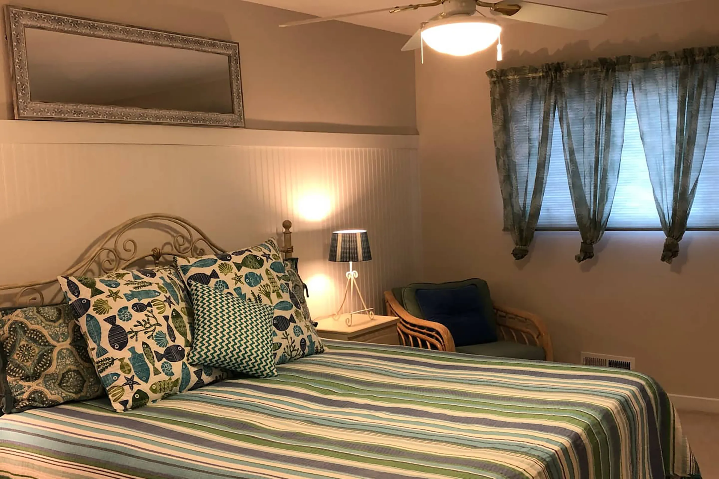 A bed with blue, green, and white striped comfortable. Next to the bed is a desk lamp and a lounge chair with wooden arms. 