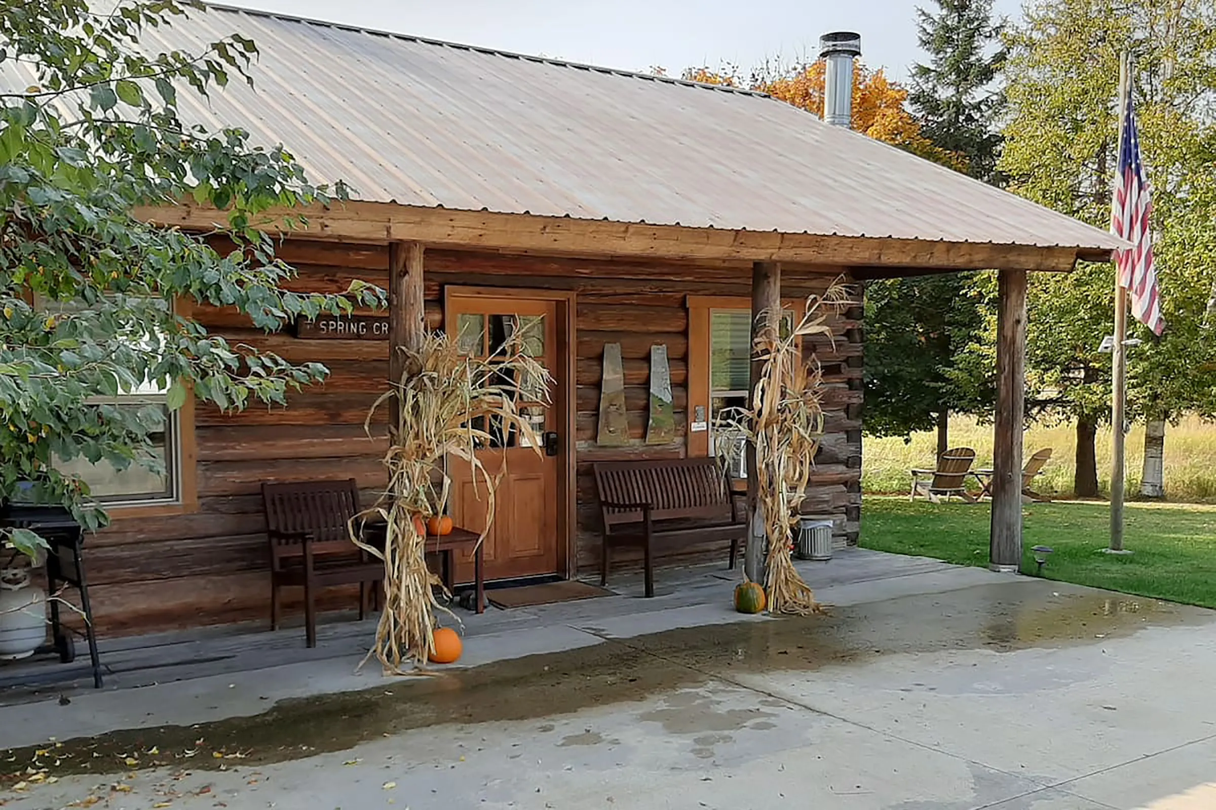 A cabin with pumpkins next to the front door and an American flag hanging from a pole to the left of the entrance to the cabin.