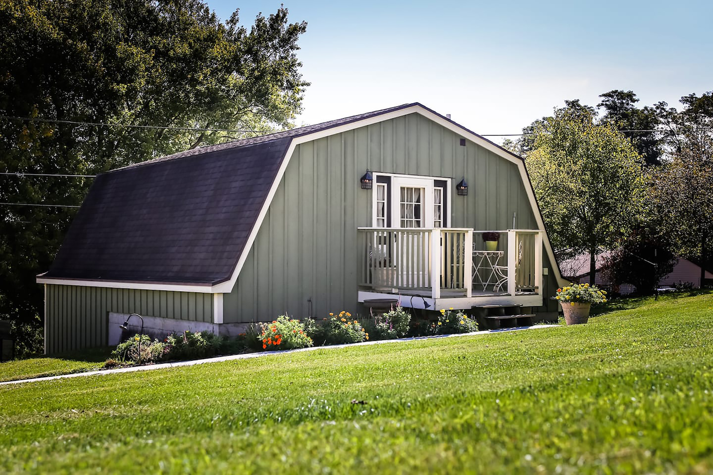 A green home with a white front door set behind a lush green yard with potted flowers.