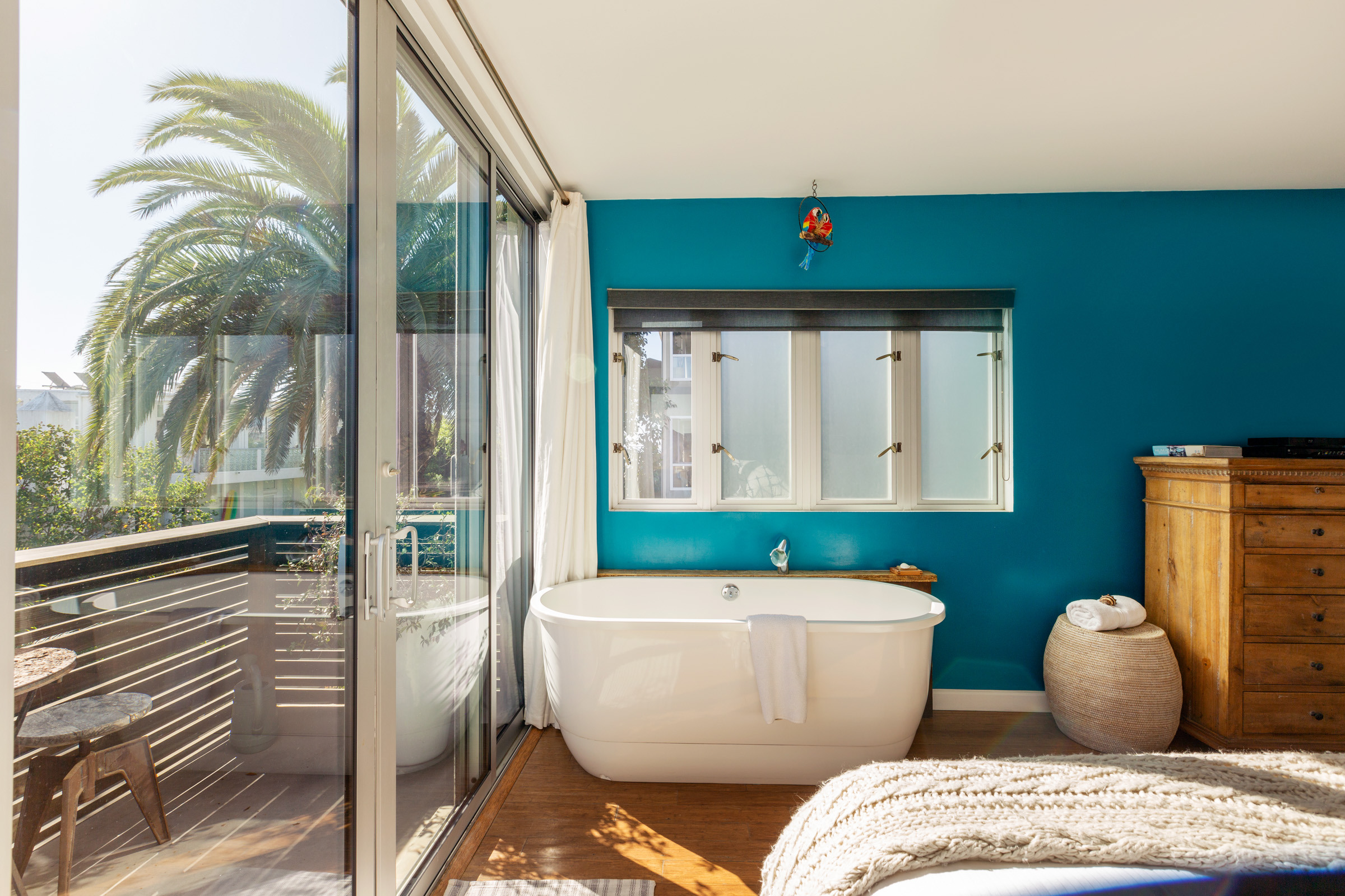 Blue painted bedroom with a bathtub against the wall with a view of palm trees and sliding glass door that leads to a balcony alongside the bedroom.