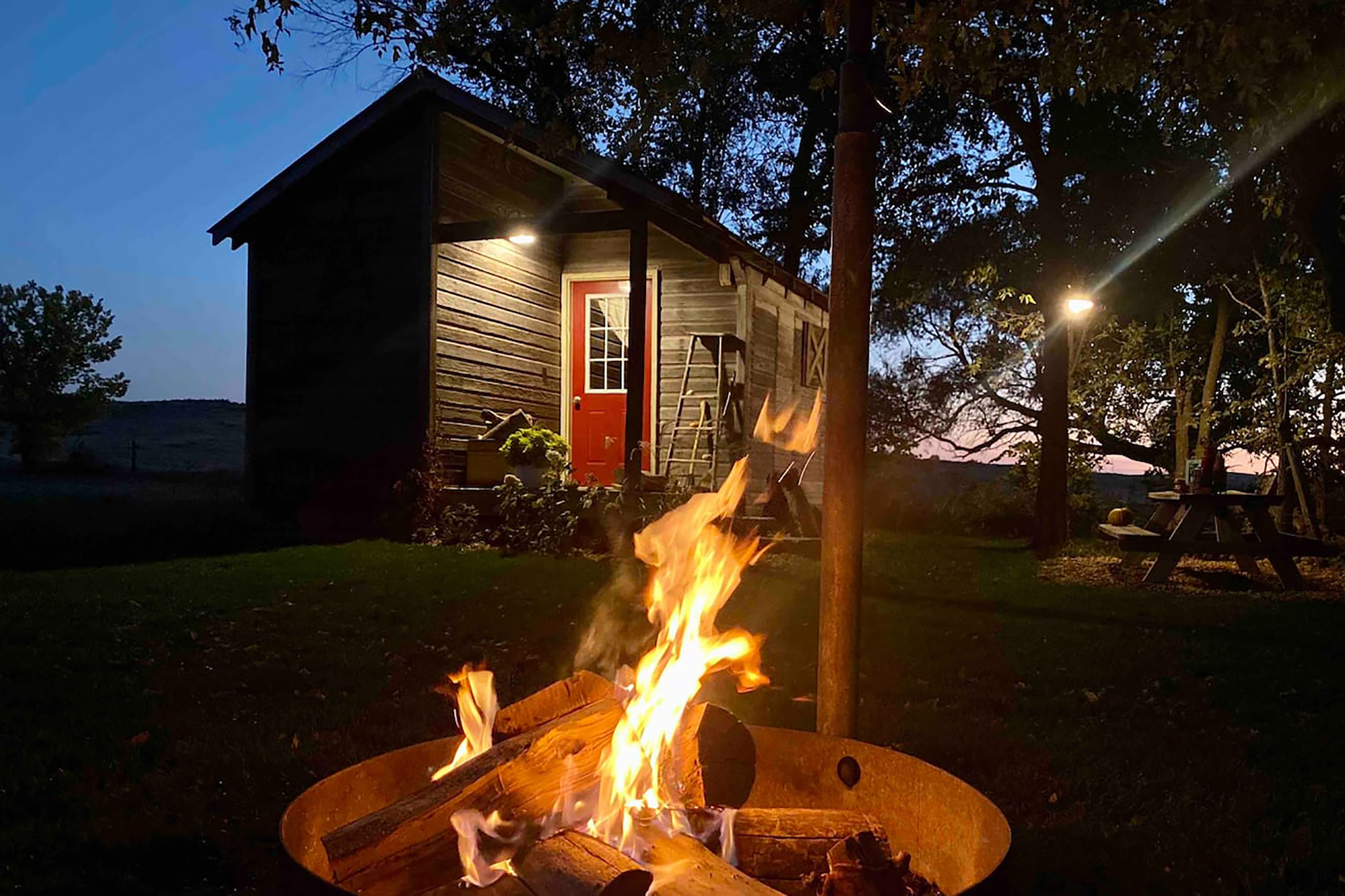 An illuminated fire pit set in front of a dark colored home with a red door.