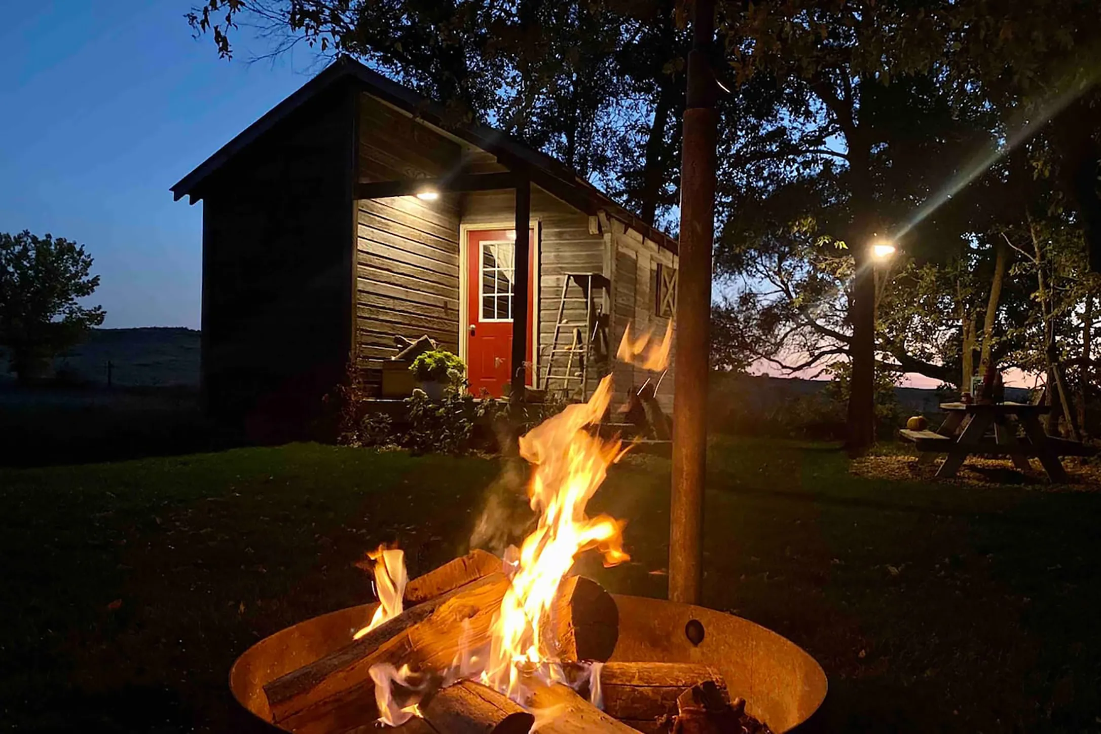 An illuminated fire pit set in front of a dark colored home with a red door. 