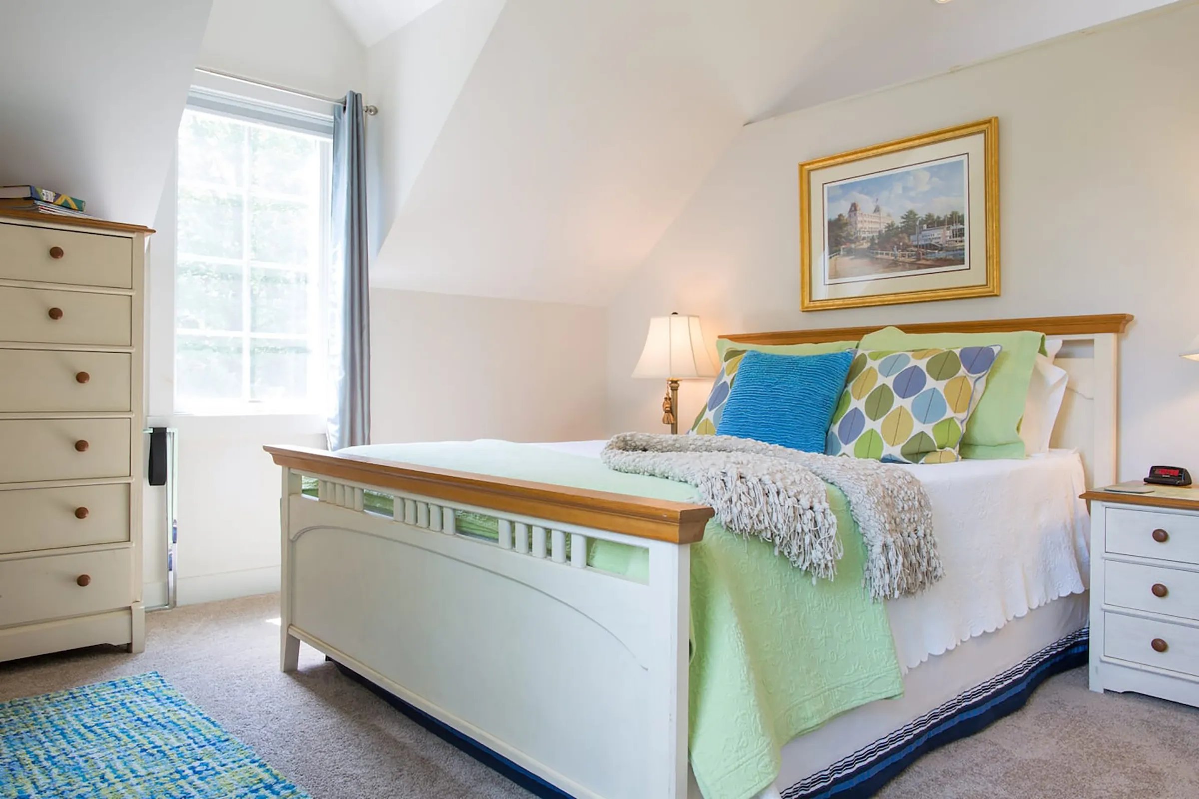 A bed with bright green and blue pillows and comforter. Above the bed hangs a picture of a building with a golden frame. 