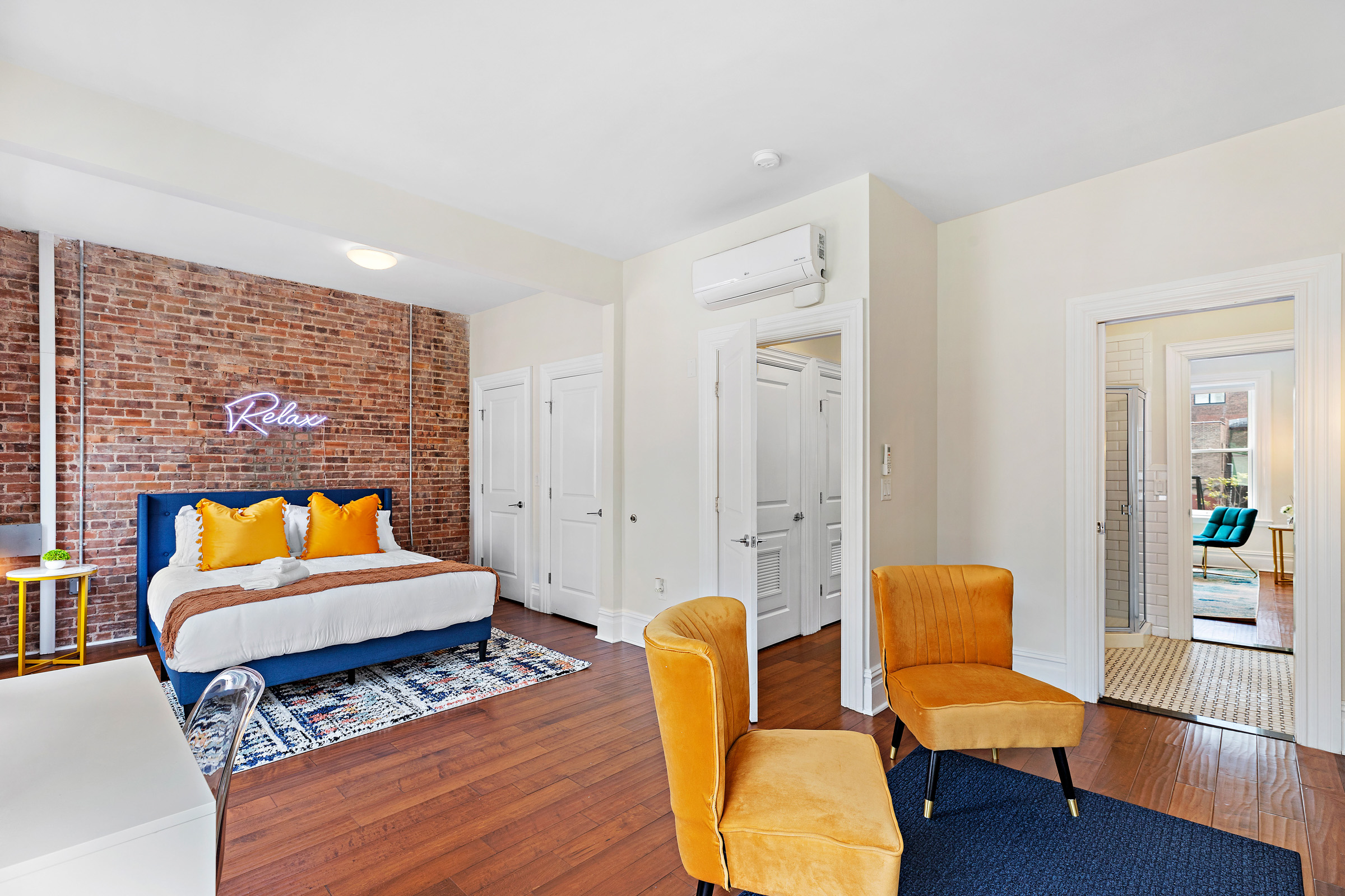 Exposed brick bedroom with orange and blue theme. Blue area rug with two orange chairs in front of a white bed with blue headboard and orange pillows.
