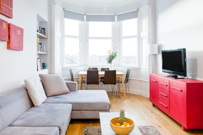 A sunlit white living room with large bay windows. A light grey sofa faces an eye-catching red TV stand.