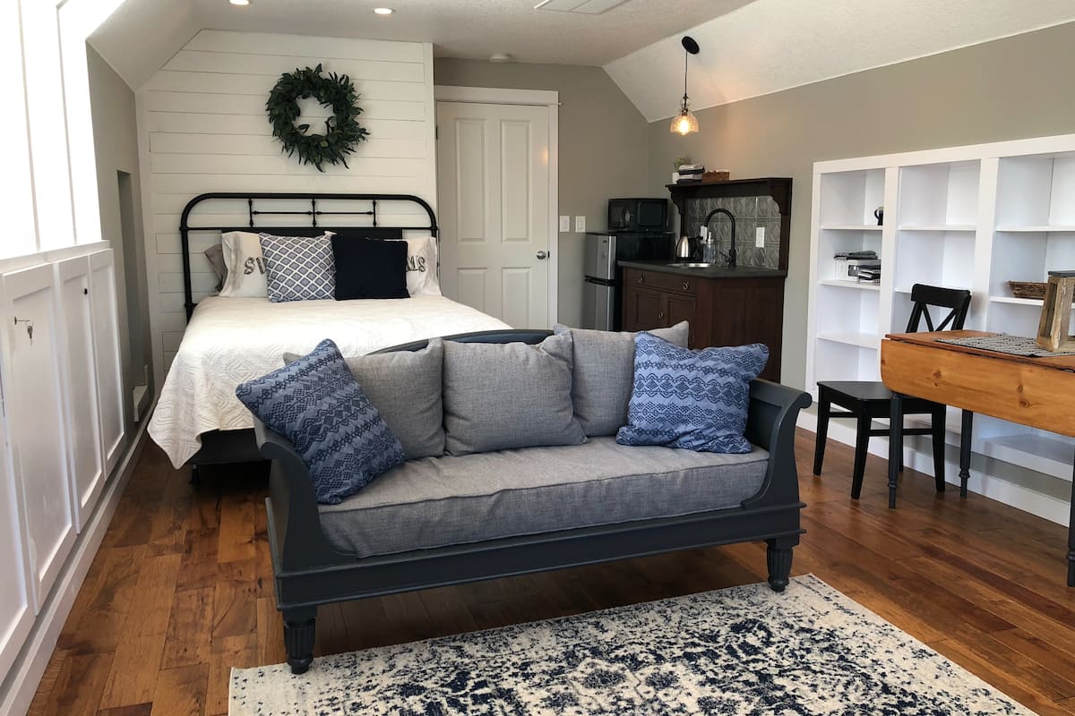 A gray colored loveseat sits in front of a bed with a white comforter.