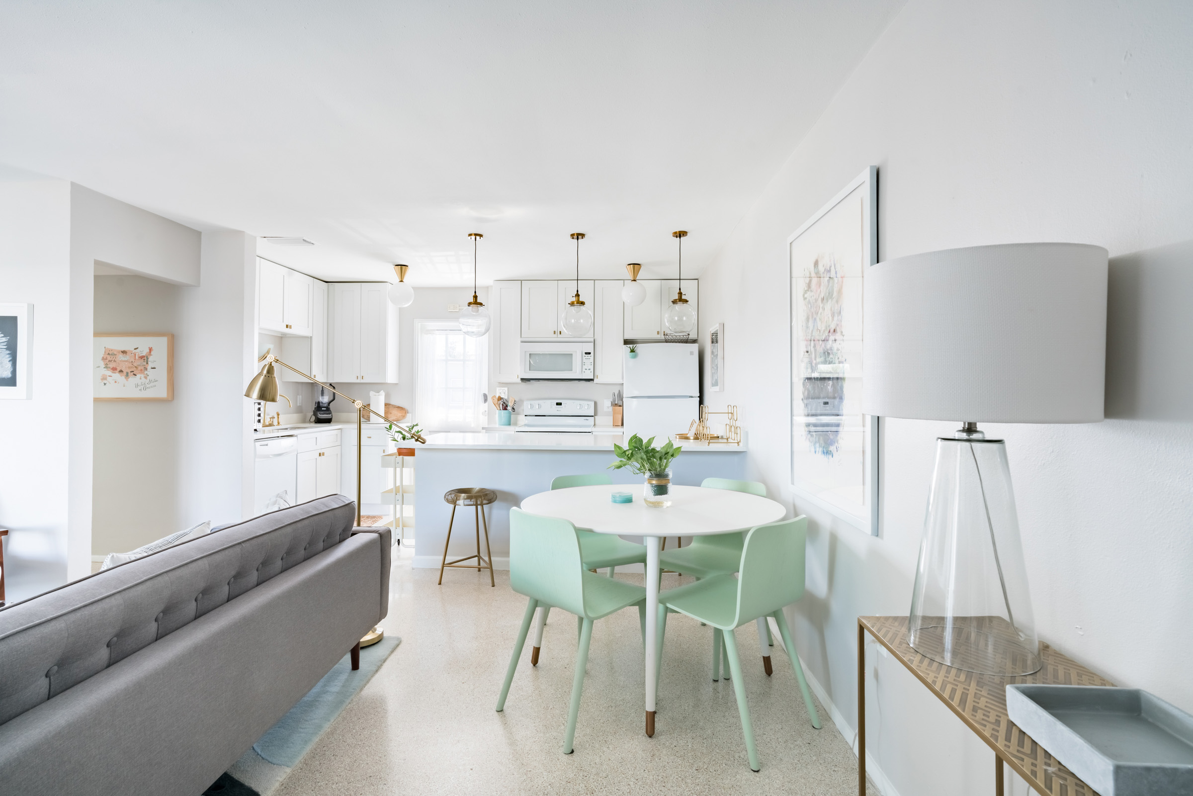 All white, open kitchen to living room scene with a mint green seating area in the foreground alongside a gray couch. Kitchenette in the background against a wall of windows letting in natural light.