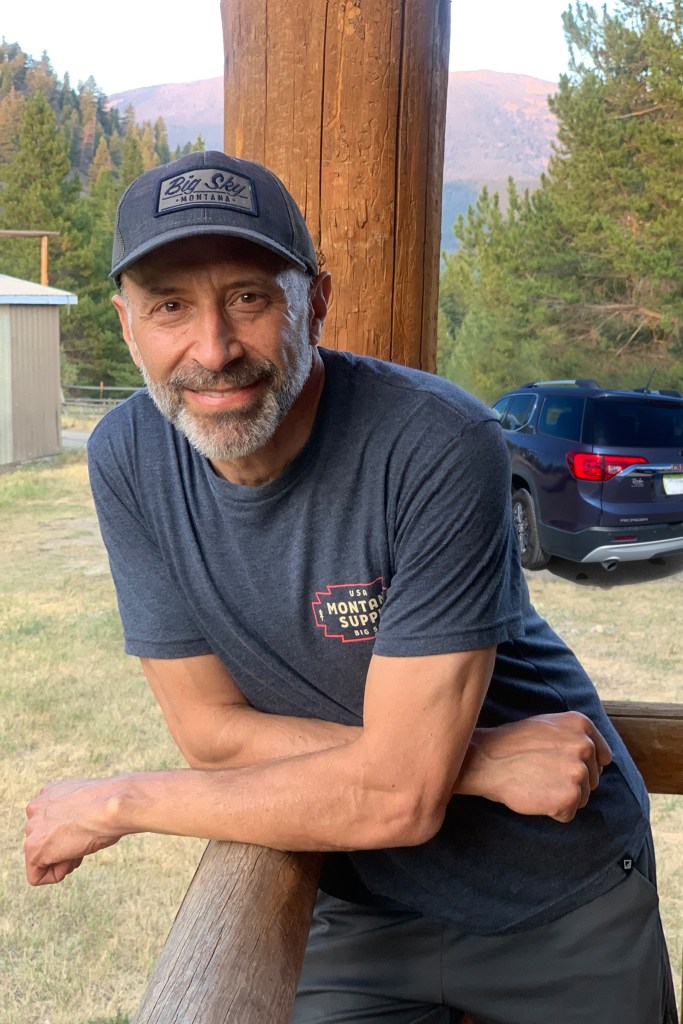 Host Paul dressed in gray t-shirt and baseball cap leaning against a wooden porch rail and his SUV in the background.