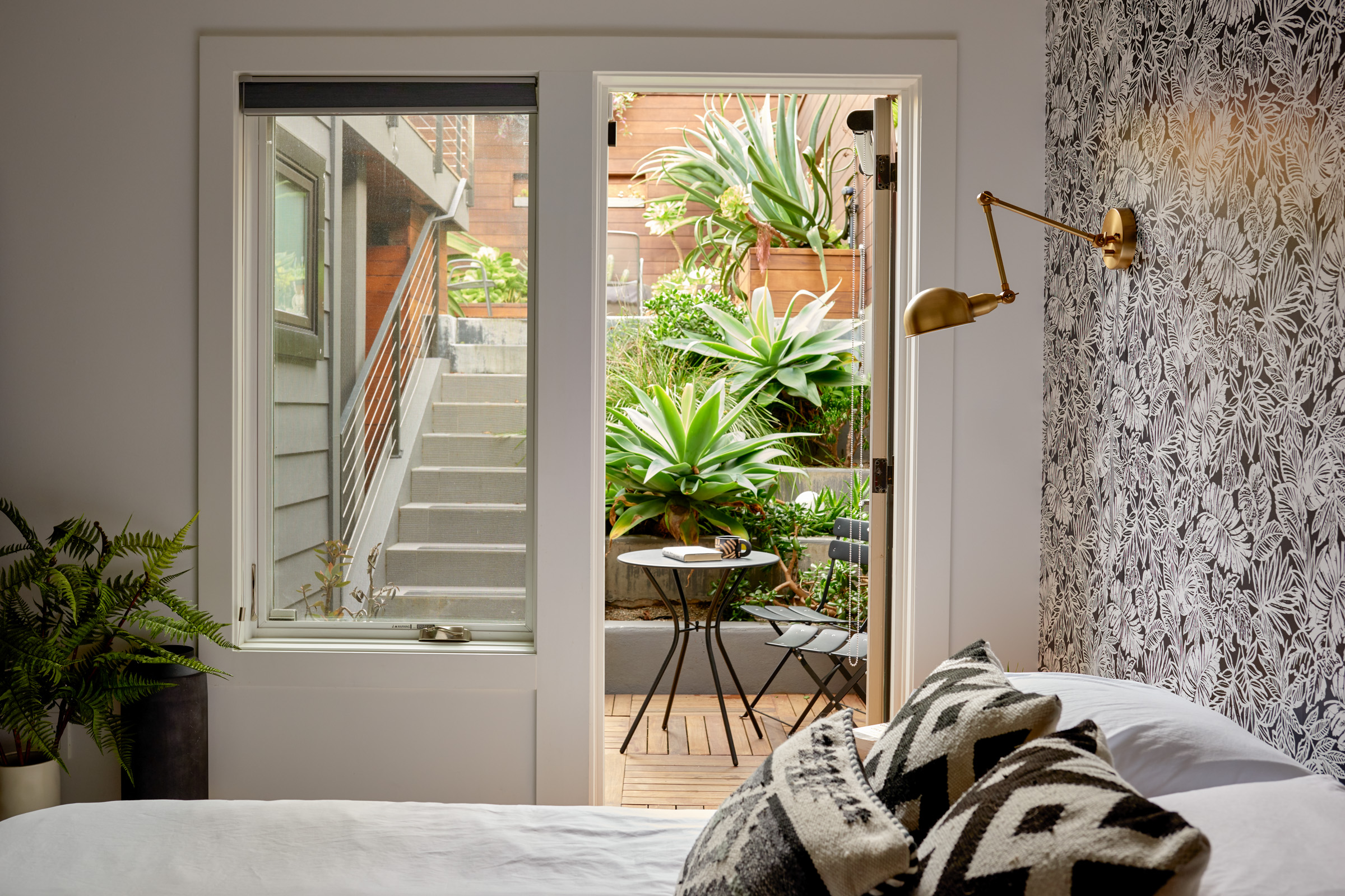 White bed with decorative pillows and gold sconce lighting above. Door in the background that leads to a set of stairs outside with greenery around them.