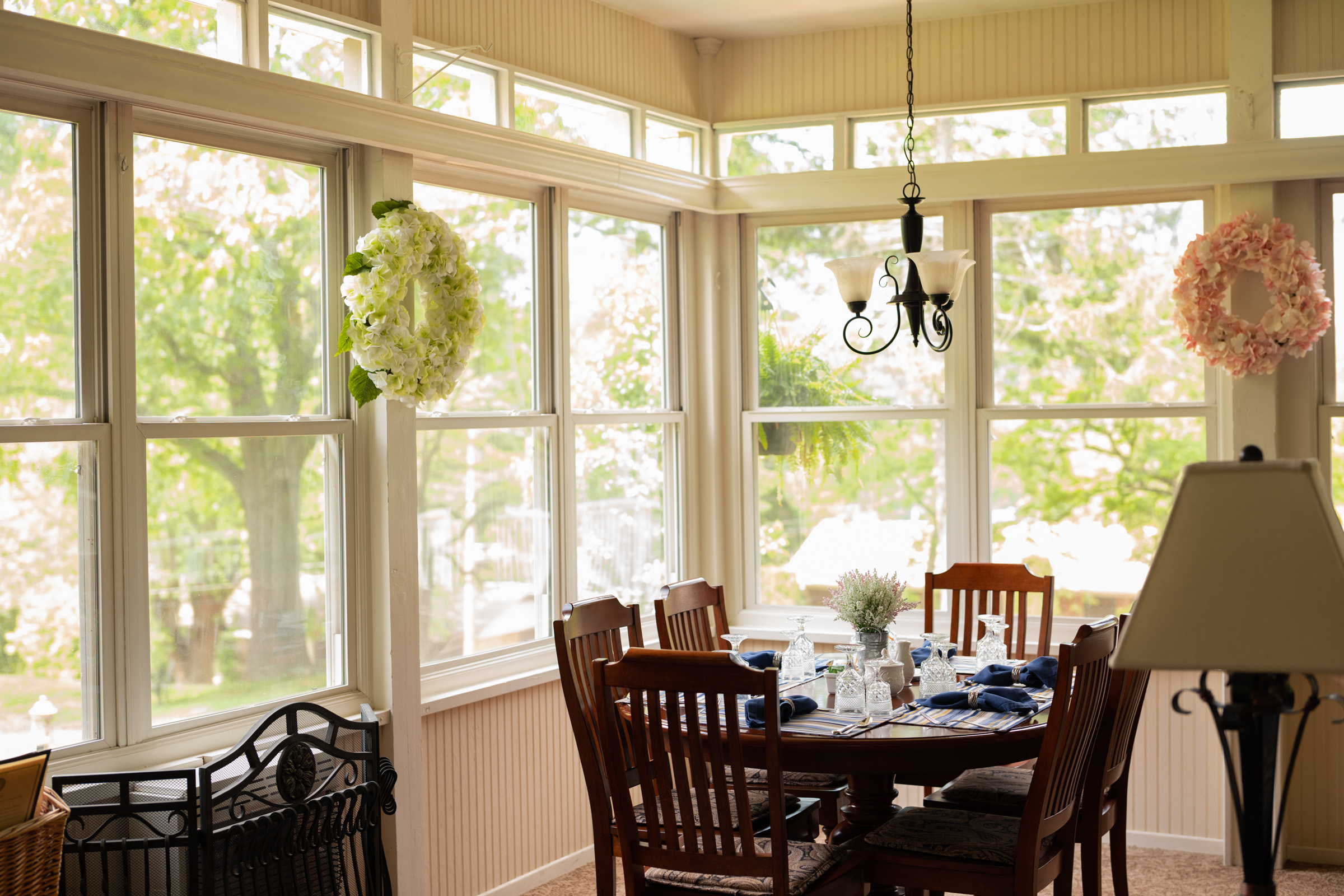 Low light dining room with windows letting in natural light. Hanging chandelier above the wooden table and chairs. You can see neighborhood trees through the windows.