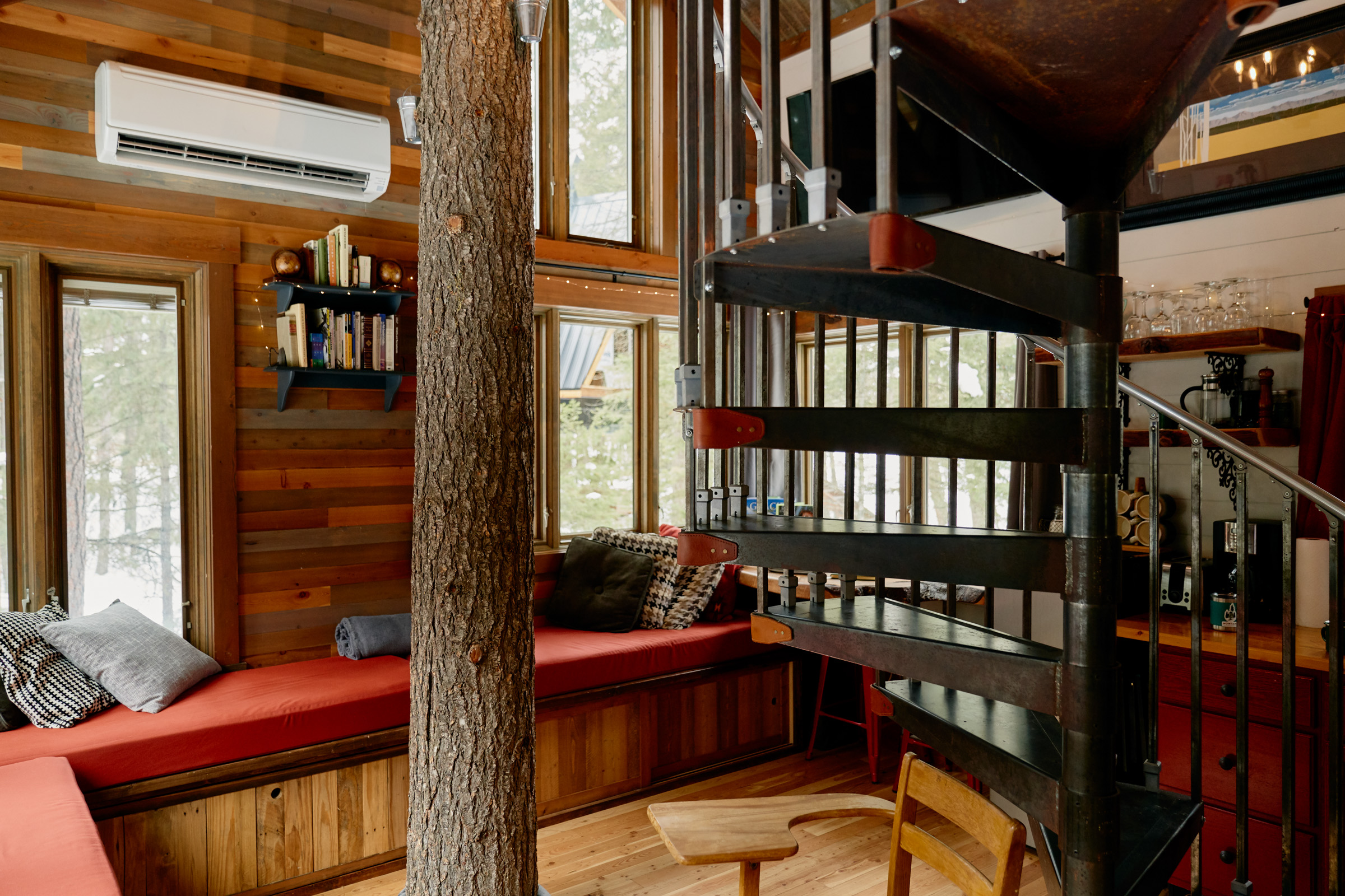 Interior log cabin with a spiral metal staircase in the foreground and a red cushioned nook in the background.