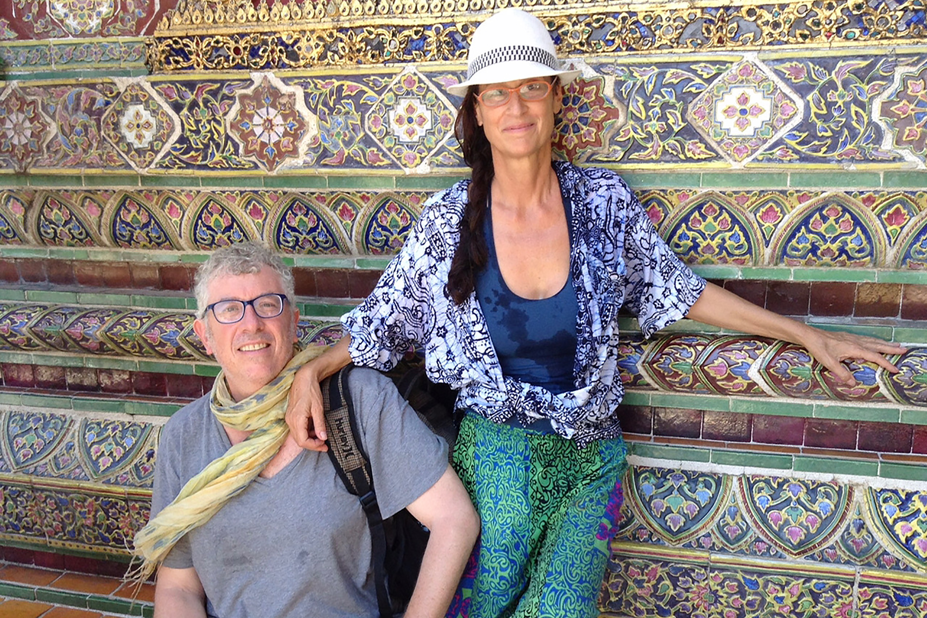 Ben and Peta standing in front of a tiled wall. Ben is wearing a gray tshirt and colorful scarf, Peta is wearing a fedora with flowy blue top and green pants.