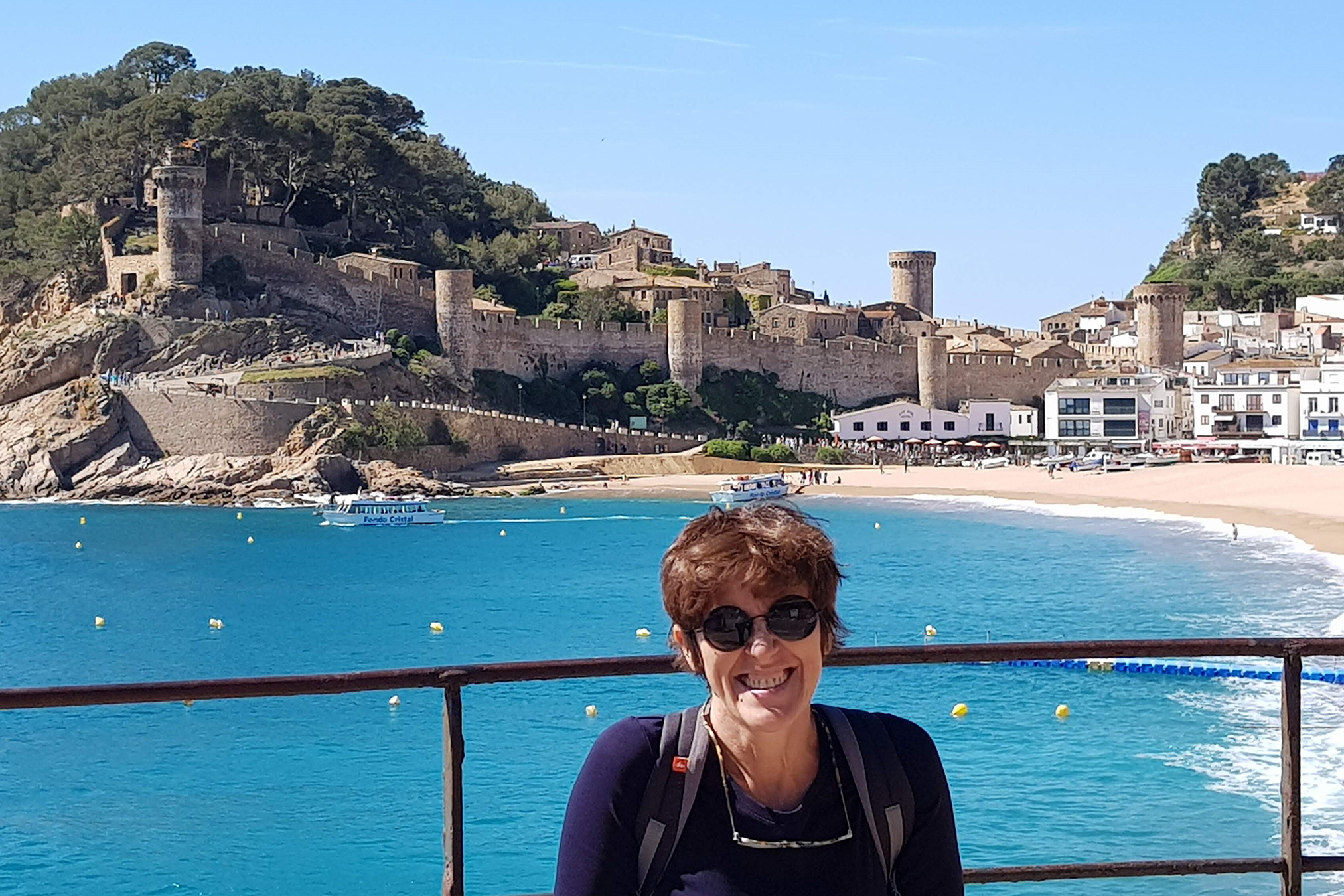 Maria in the foreground wearing a black jacket and sunglasses. Behind her you see crystal blue waters and a beachtown.