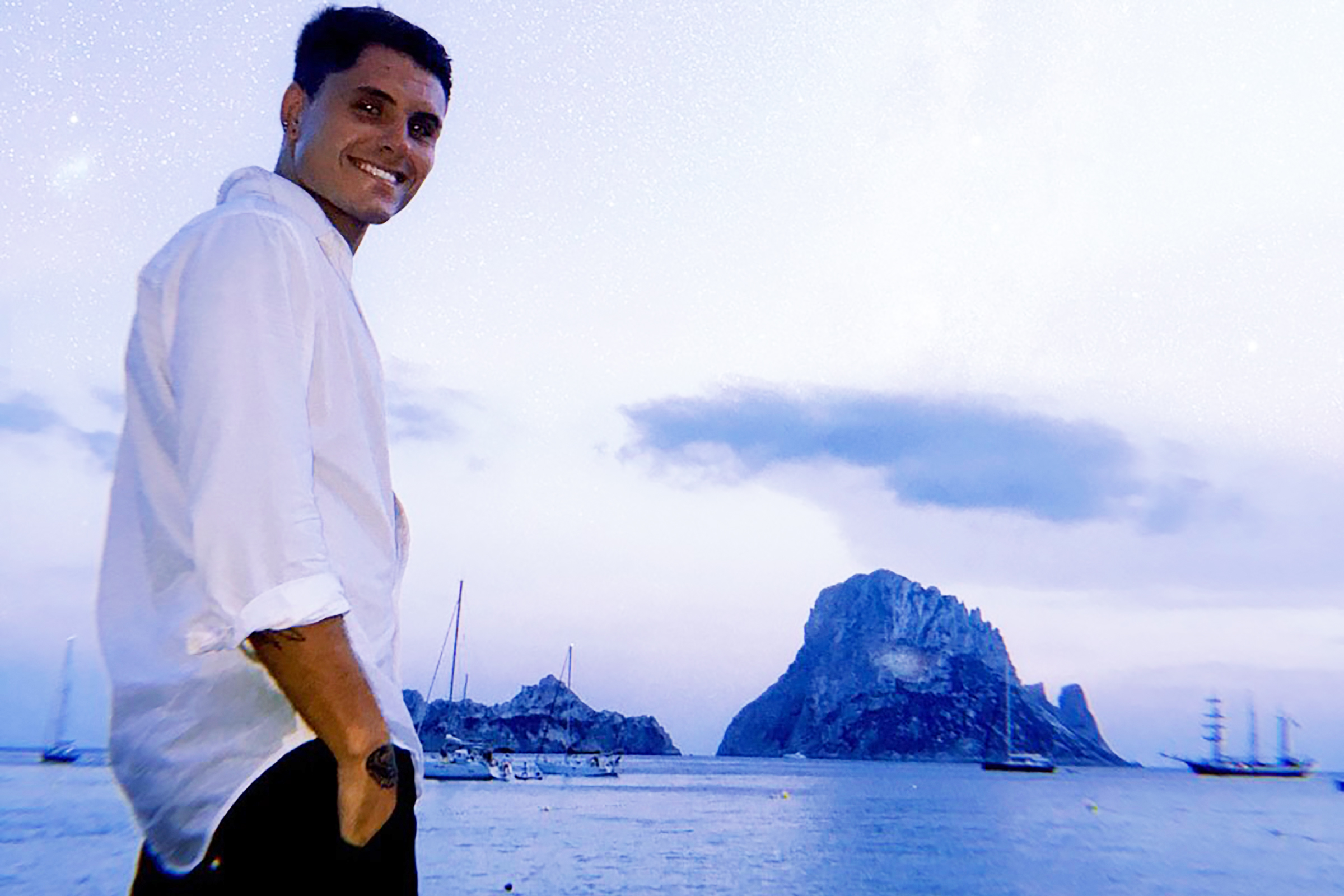 James B. standing on the beach dressed in a white shirt and dark pants with sailboats and a large rock mass on the water in the background. Very purple-y night sky.