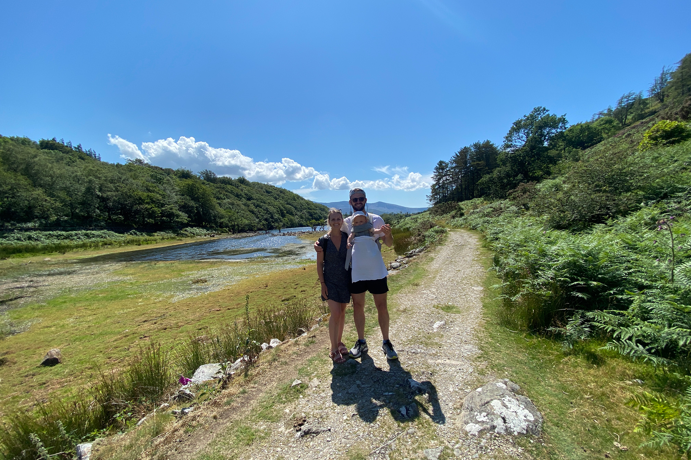 James G. with his fiance and baby standing on a trail surrounded by green grass and a forest.