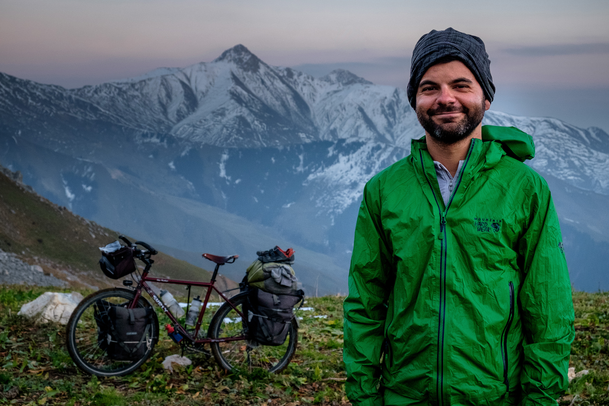 Jonathan standing in a beanie and green windbreaker with his bicycle and a mountain in the background.