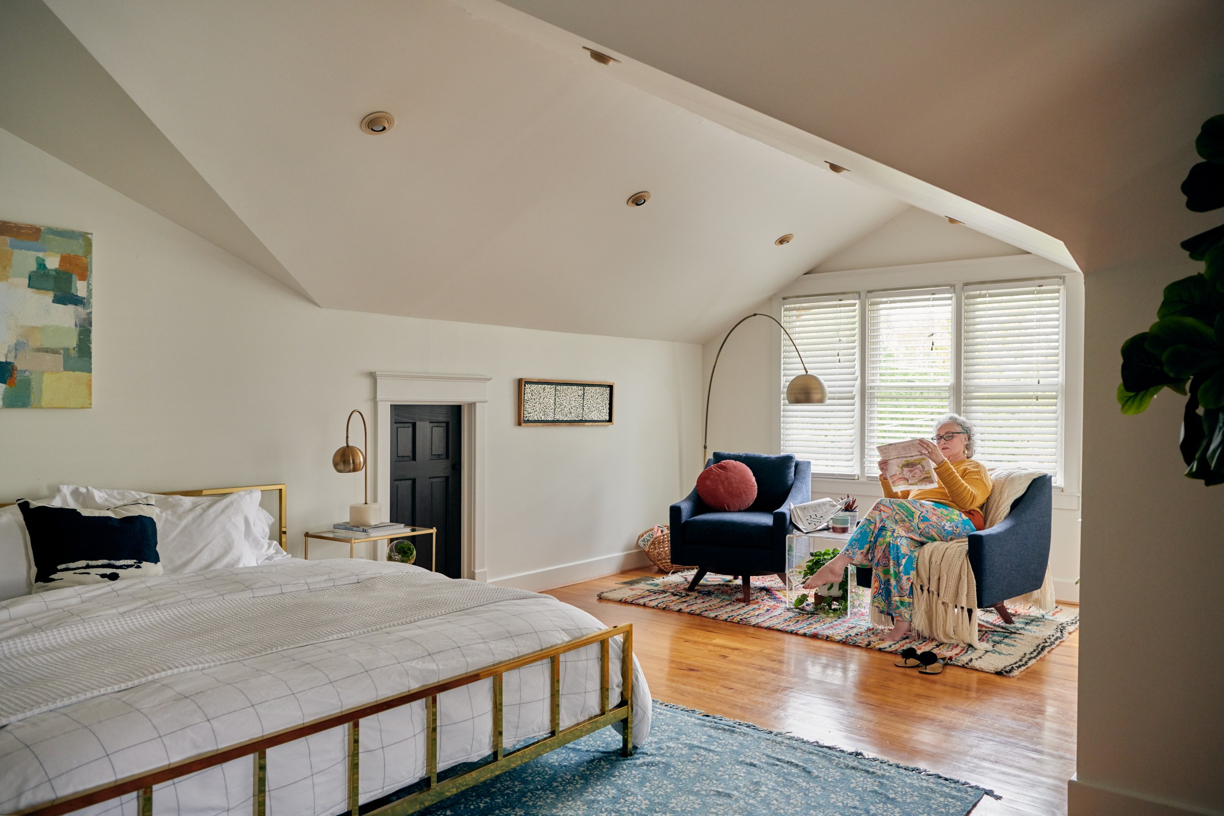 An older woman wearing a yellow shirt and blue jeans, sitting on a blue chair in a bedroom, reading a newspaper