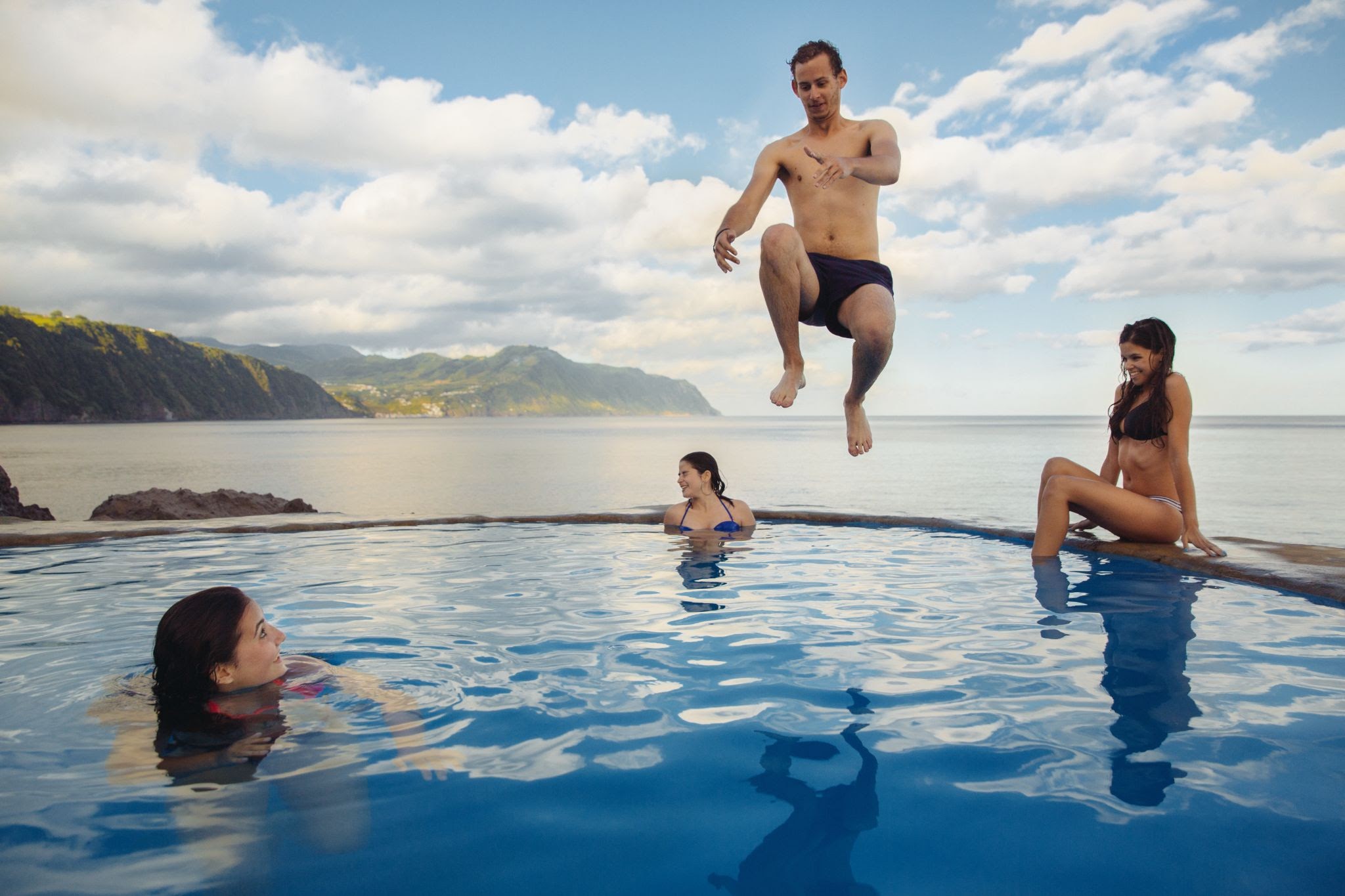 A young person jumps into an infinity pool while others watch excitedly.
