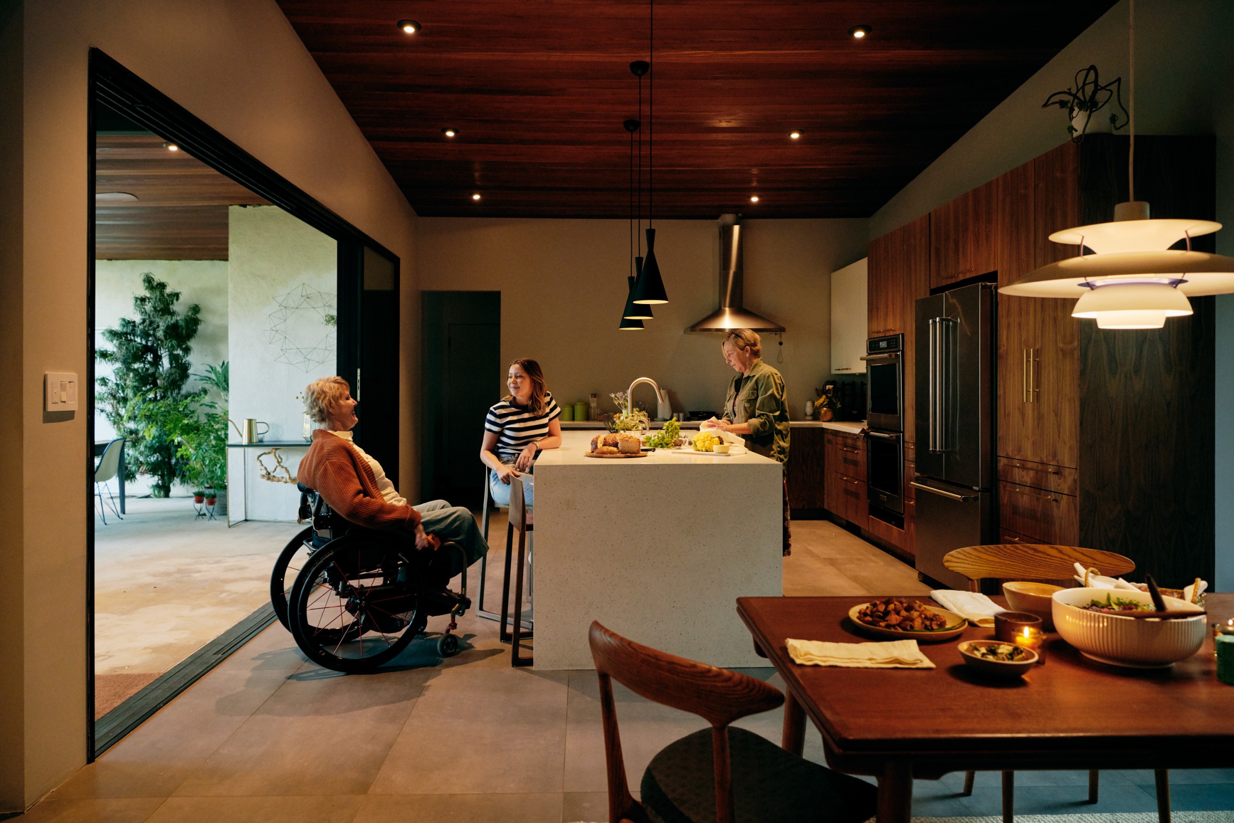 A person in a wheelchair having a conversation with two fellow travelers in an open floor plan kitchen at their Airbnb. Dining table set for a meal is in the foreground.
